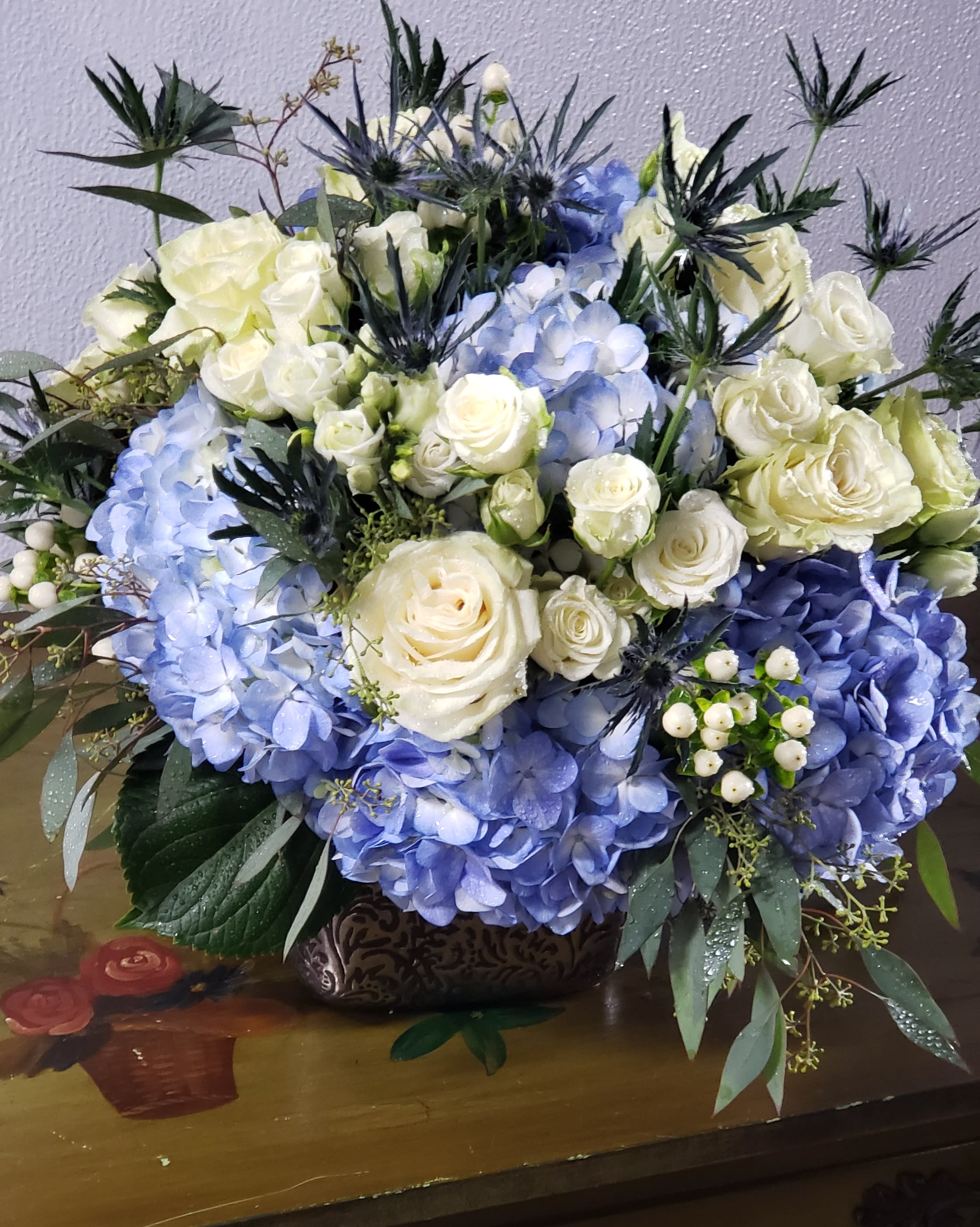 Lush and Layered Blues  -  Elegant copper cube, filled with perry winkle blue hydrangea blooms,white roses,  spray roses, complimented with white hypericum berries,eucalyptus and blue thistle . This is all the inspiration you need for your holiday table or a great gift for a special hostess! *container may vary*