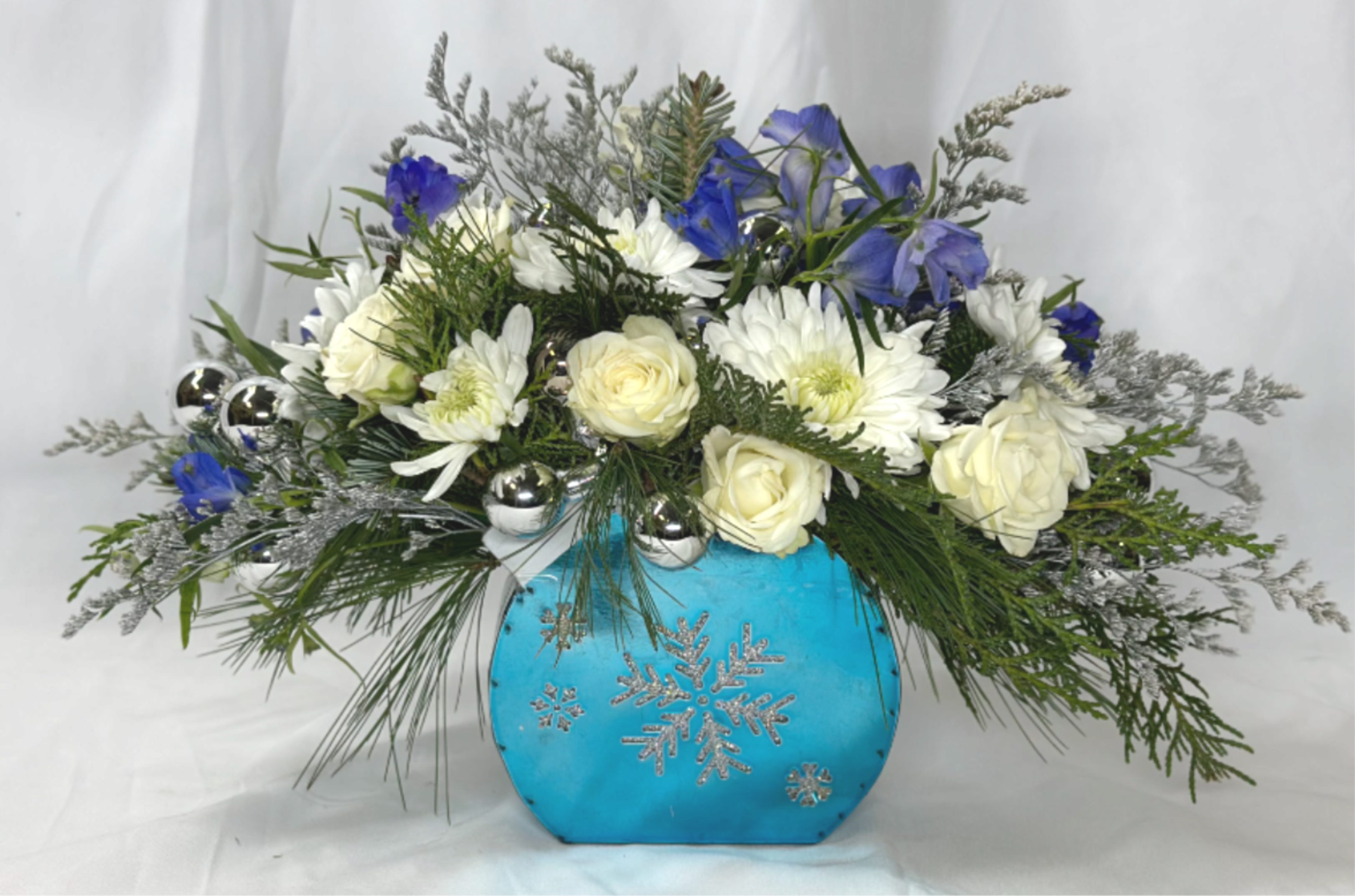 Holiday Blues - This cheerful blue and white arrangement, featuring spay roses, cushion mums, and delphinium, is sure to chase away the winter blues. The bright silver decoration will be enjoyed at Hanukkah, Christmas, or any Winter event or holiday. This is a Leary's Flowers exclusive design. If featured flowers are unavailable will will be sure to substitute other beautiful flowers of equal or greater value