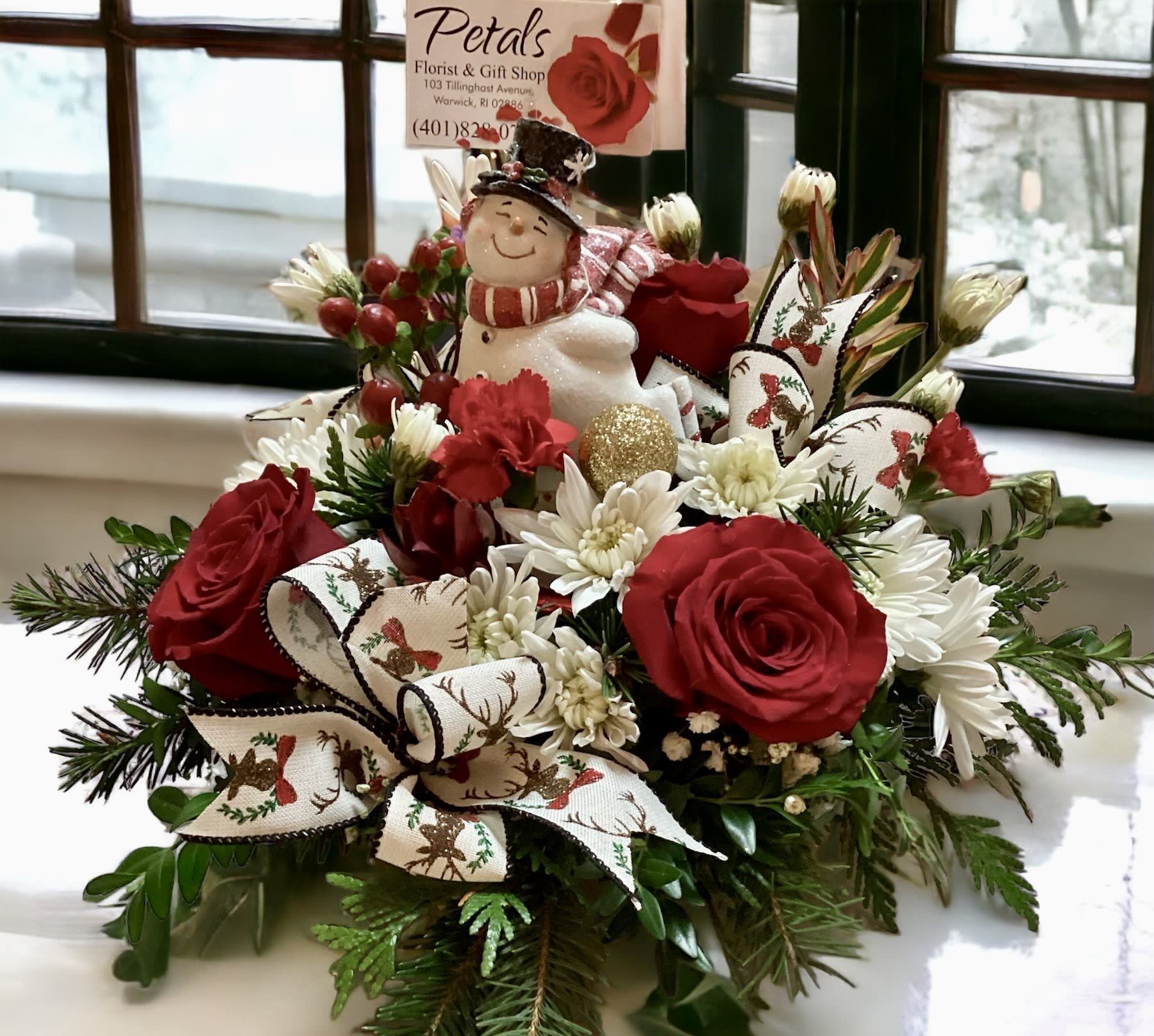 Shimmering Snowman Centerpiece by Petals - Introducing the cherished Shimmering Skating Snowman arrangement.  This limited-edition piece is designed to be a gift that keeps giving, offering joy for years to come. The snowman figure is elegantly displayed within a low bowl arrangement, featuring the delightful, sweet-scented winter greens, festive red roses, white chrysanthemums, hypericum berries, and charming fabric bows (designs may vary). This is a perfect, ready-to-display gift for anyone on your list. As this is a limited item, please pre-order to secure yours today. Offer valid only while supplies last. Call Cheryl at 401-828-0791 with any inquiries.