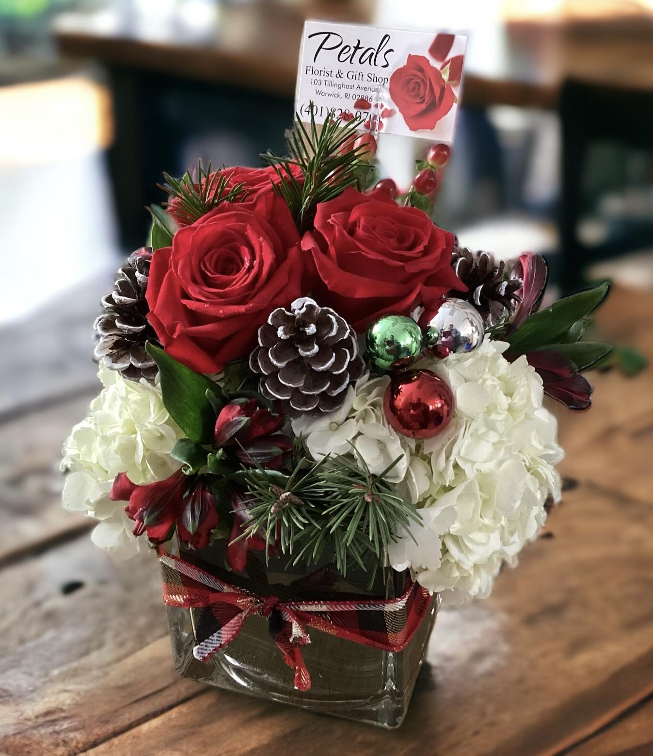 Cozy Christmas Cube by Petals -  This holiday season, delight someone special with the ultimate expression of Christmas coziness: our exquisite Cozy Christmas Cube arrangement. Meticulously designed to bring the fresh, invigorating scents of the season indoors, this stunning piece features a lush blend of fresh winter greens, velvety red roses, soft white hydrangeas, delicate alstroemeria, festive hypericum berries, natural pine cones, and elegant glass balls. Each element is artfully nestled within a premium 5x5x5-inch glass cube vase, beautifully finished with a charming check wrap bow (pattern may vary). It’s the perfect, ready-to-gift centerpiece for any home and anyone on your list this year. Spread joy and the true spirit of Christmas with this simply delightful creation!