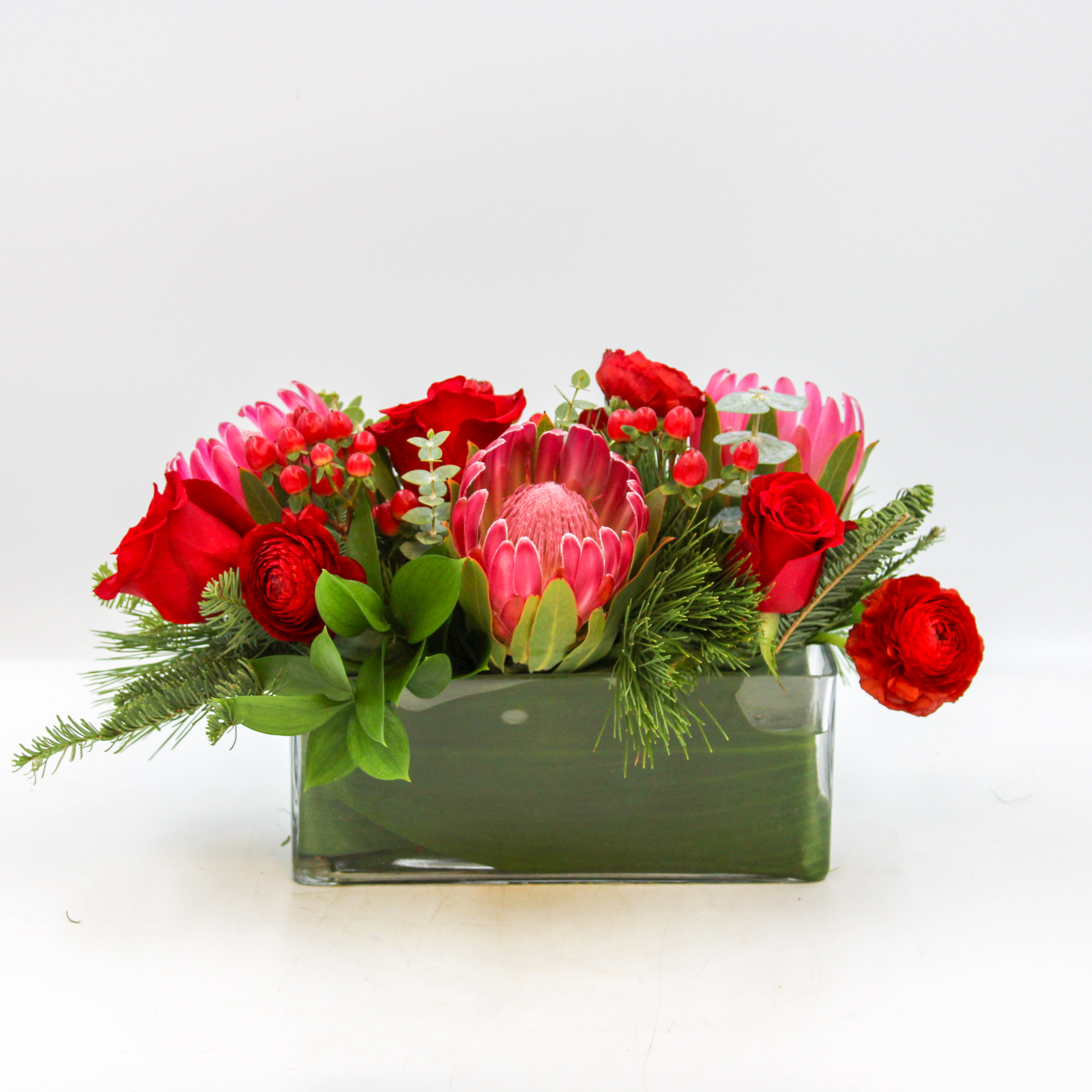 Prancer - Prancer is a luxurious centerpiece with beautiful Red Protea, Red Roses, Red Ranunculus, Red Hypericum Berries, Eucalyptus and mixed evergreens arranged in a leaf lined glass rectangular vase. Prancer is approximately 13 inches in length. 