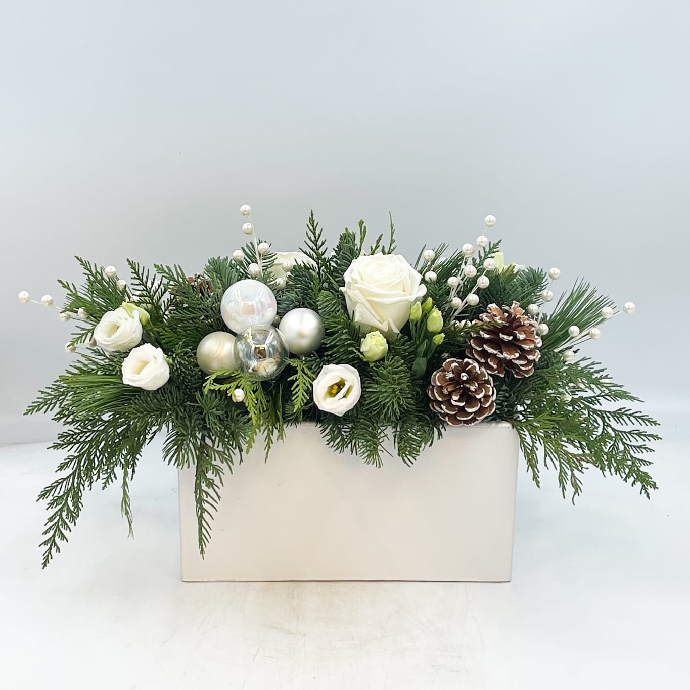 White Christmas - A beautiful gold and white Christmas Centerpiece, Christmas Dreaming features Snowcapped Pinecones, Mixed White and Iridescent Ornaments, Pearl Berry stems, White Lisianthus and White Roses in a bed of mixed evergreens arranged in a keepsake White Ceramic Planter.