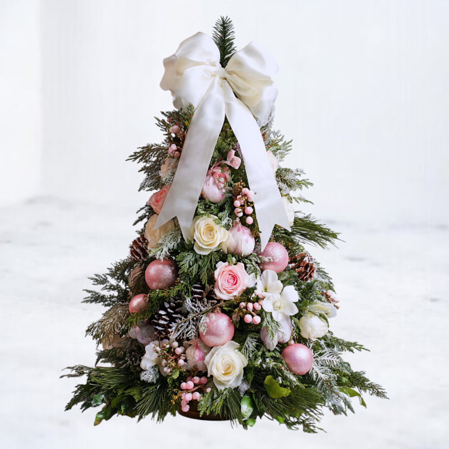 Sugarblush Winter Tree - Soft, romantic, and winter-kissed, the Sugarblush Winter Tree blends pink roses and white spray roses, frosted evergreens, pinecones, and shimmering pink ornaments for a dreamy, snow-touched holiday aesthetic. Topped with a silky ivory bow, this handcrafted tabletop tree brings a delicate, ballet-inspired charm to any space.  Ideal for mantels, kitchen islands, nursery décor, holiday gifting, or adding a refined feminine touch to seasonal styling. Each tree is built by Whiskey Wallflower using fresh winter greens and premium accents for a full, layered, luxurious look.  Sizes: Standard: Approximately 12 inches tall Deluxe: Approximately 15–16 inches tall Premium: Approximately 18–20 inches tall (Premium size shown in photo)  Available for same-day holiday and Christmas delivery in Magnolia, The Woodlands, Conroe, Tomball, and surrounding Houston areas.