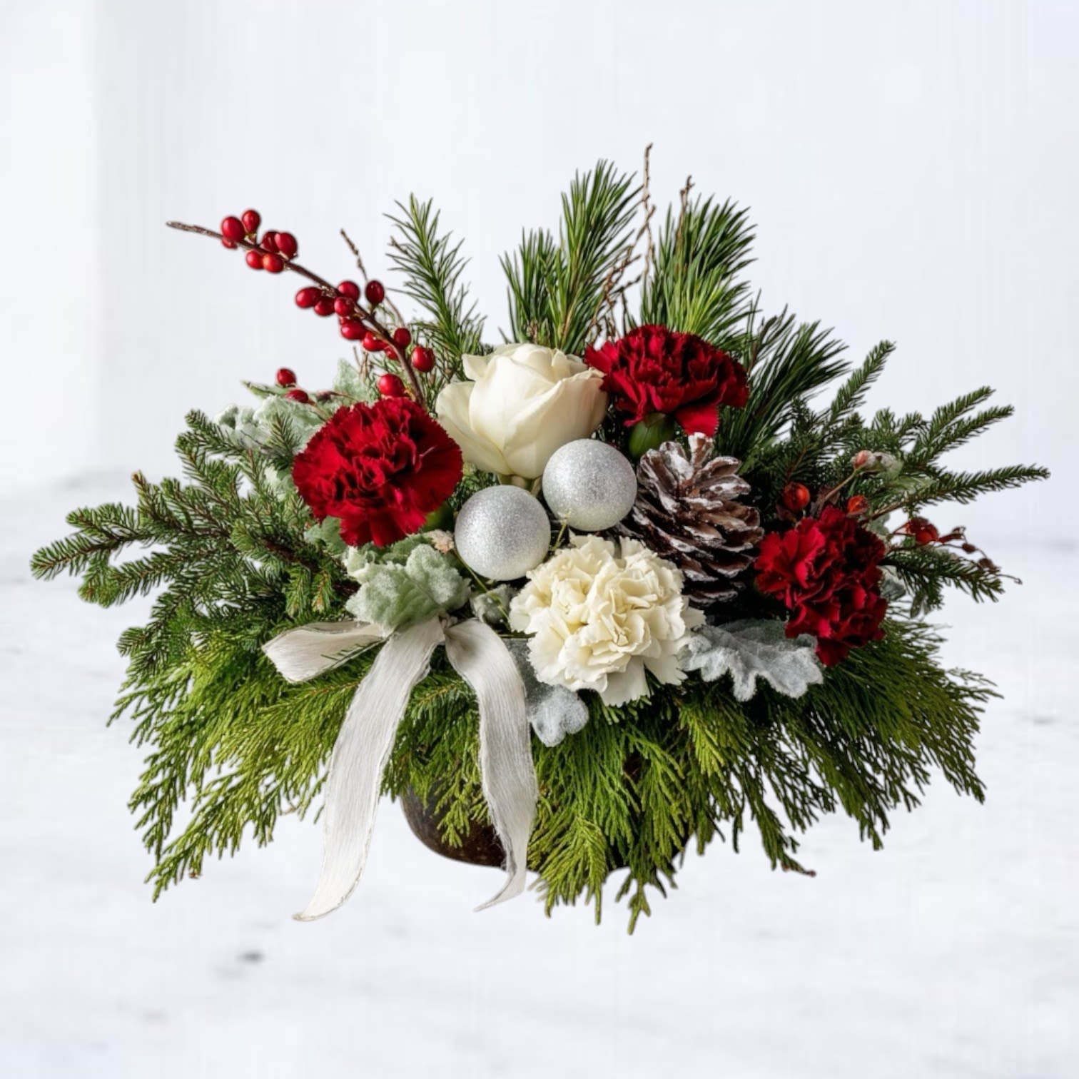 Snowberry Pine - The Snowberry Pine Arrangement blends classic holiday charm with soft winter texture. Featuring red carnations, ivory roses, snow-kissed lamb’s ear, natural pinecones, silver ornaments, and fresh evergreens, this handcrafted design brings warmth, fragrance, and festive color to any space.  Perfect for holiday tables, kitchen islands, office desks, or seasonal gifting, the Snowberry Pine Arrangement offers a cozy, polished Christmas aesthetic without overwhelming smaller areas. Designed by Whiskey Wallflower, each piece is built with premium winter greens and long-lasting blooms for a full, layered finish.  Available for same-day Christmas and holiday delivery in Magnolia, The Woodlands, Conroe, Tomball, and surrounding Houston areas.