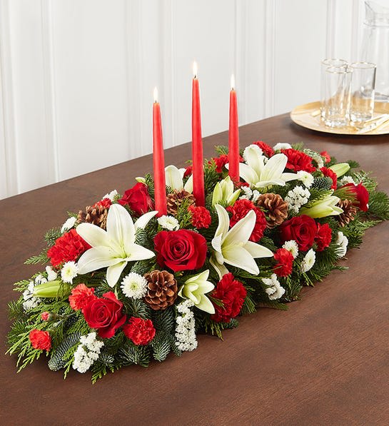 Holiday Happiness Traditional Centerpiece - Our traditional table arrangement creates the perfect centerpiece for your Christmas celebration. Beautifully designed with long-lasting blooms in seasonal colors, this fragrant gathering features red taper candles to cast a warm, cozy glow throughout the holidays.  Centerpiece arrangement with red roses, carnations and mini carnations; white Asiatic lilies, button poms and statice; accented with assorted Christmas greenery and pinecone picks