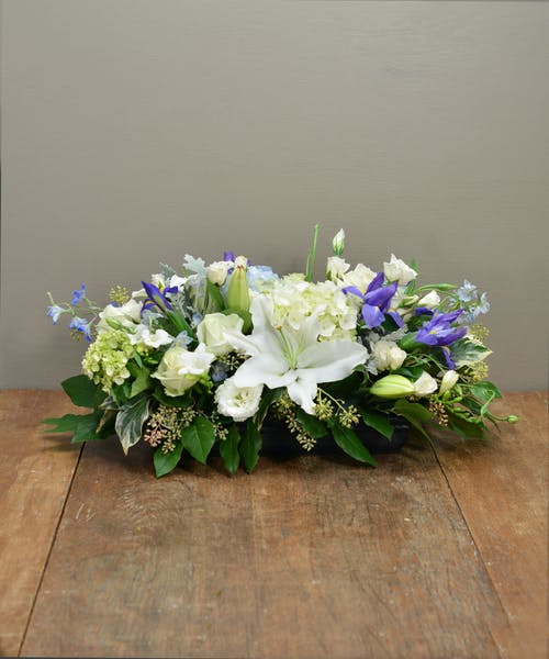 Traditional holiday centerpiece in blue and white - This is a sophisticated, long low centerpiece in complementary hues of blue and white. This lovely creation is made with blue delphinium, hydrangea, iris, lisanthus, white roses, white lilies and seasonal greens It would look lovely on any holiday table. 