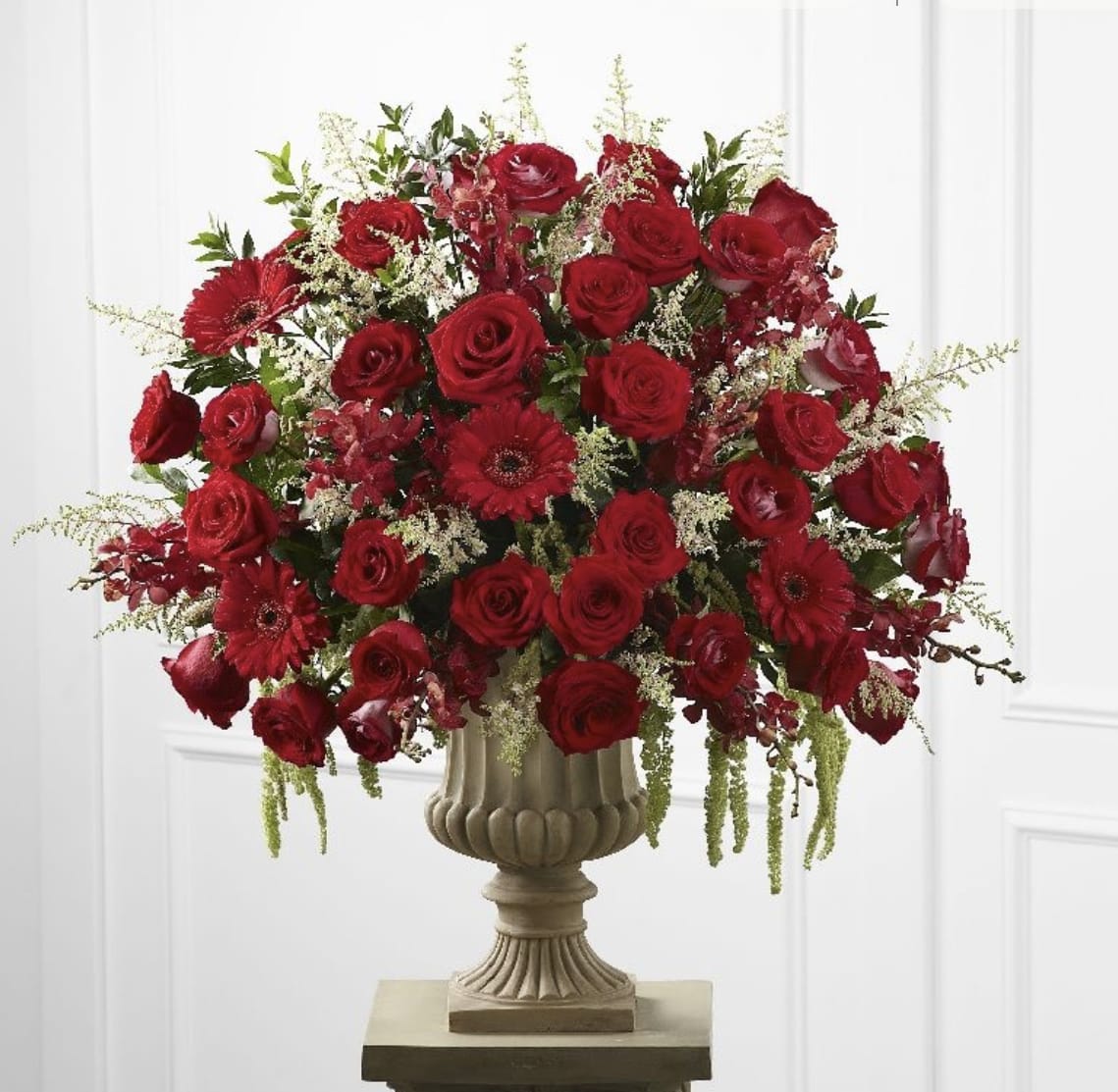 Red Majesty Memory pre-order Day before - Rich, majestic, and deeply heartfelt, this arrangement is powerful expression of love and remembrance. Designed in a classic stone vase, this arrangement of red roses, gerbera daisies, and seasonal blooms- each flower chosen to symbolize devotion, respect, and enduring memory.  Soft touches of cascading greenery and delicate filler blooms add movement and grace, creating a full, radiant display that commands presence without overwhelming the space. This arrangement is ideal for funeral services, memorial gatherings, or as a family tribute piece- honoring a life that was cherished, respected, and forever loved.