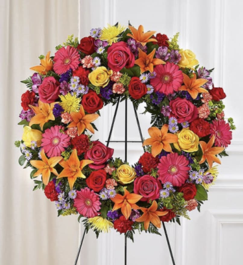 Bright Memories Wreath - Bold, uplifting, and filled with color, the Bright Memories Wreath honors a life lived with joy and light. This radiant design blends roses, lilies, gerbera daisies, and seasonal blooms in shades of orange, fuchsia, yellow, and red- creating warm, celebratory expression of love.  This circular shape symbolizes eternity and the unending bond shared with those we cherish. Each flower is placed with intention, forming a wreath that feels both vibrant and heartfelt- a tribute that celebrates personality, passion, and the beautiful moments remembered forever.  Perfect for funeral services, memorials, or celebration-of-life ceremonies, this wreath brings comfort through color and honors a life that continues to shine.