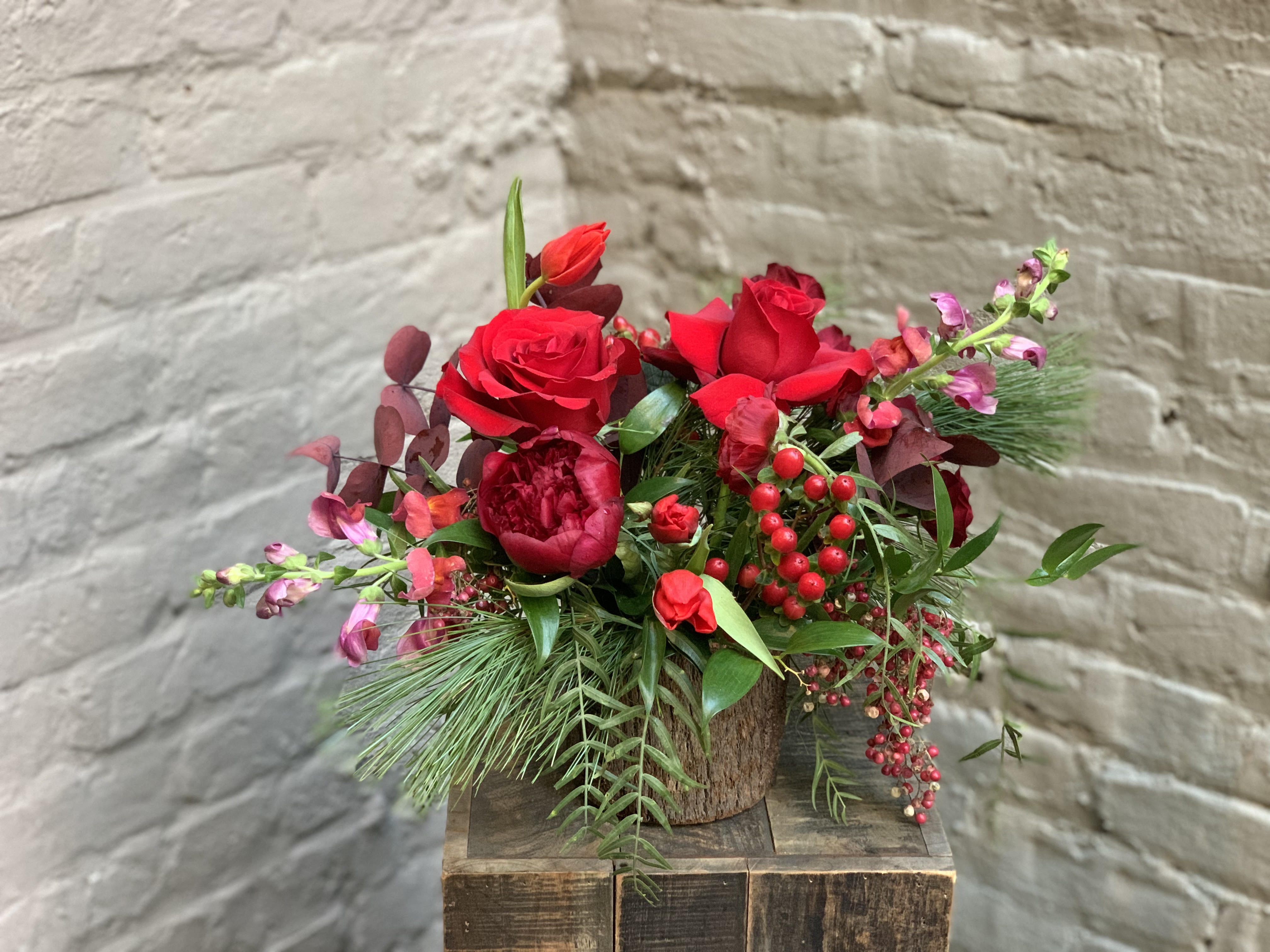 Cozy Little Christmas - Curl up by the fireplace, and make the hot cocoa -- it's time to step into a cozy little Christmas time!  This arrangement features a rich palette of red blooms; shades of candy apple, merlot, cinnamon, and luxardo cherry.  Fresh pepperberry and pine adorn this woodsy arrangement and whisper winter wishes to you! Current vessel is a clear glass vase.