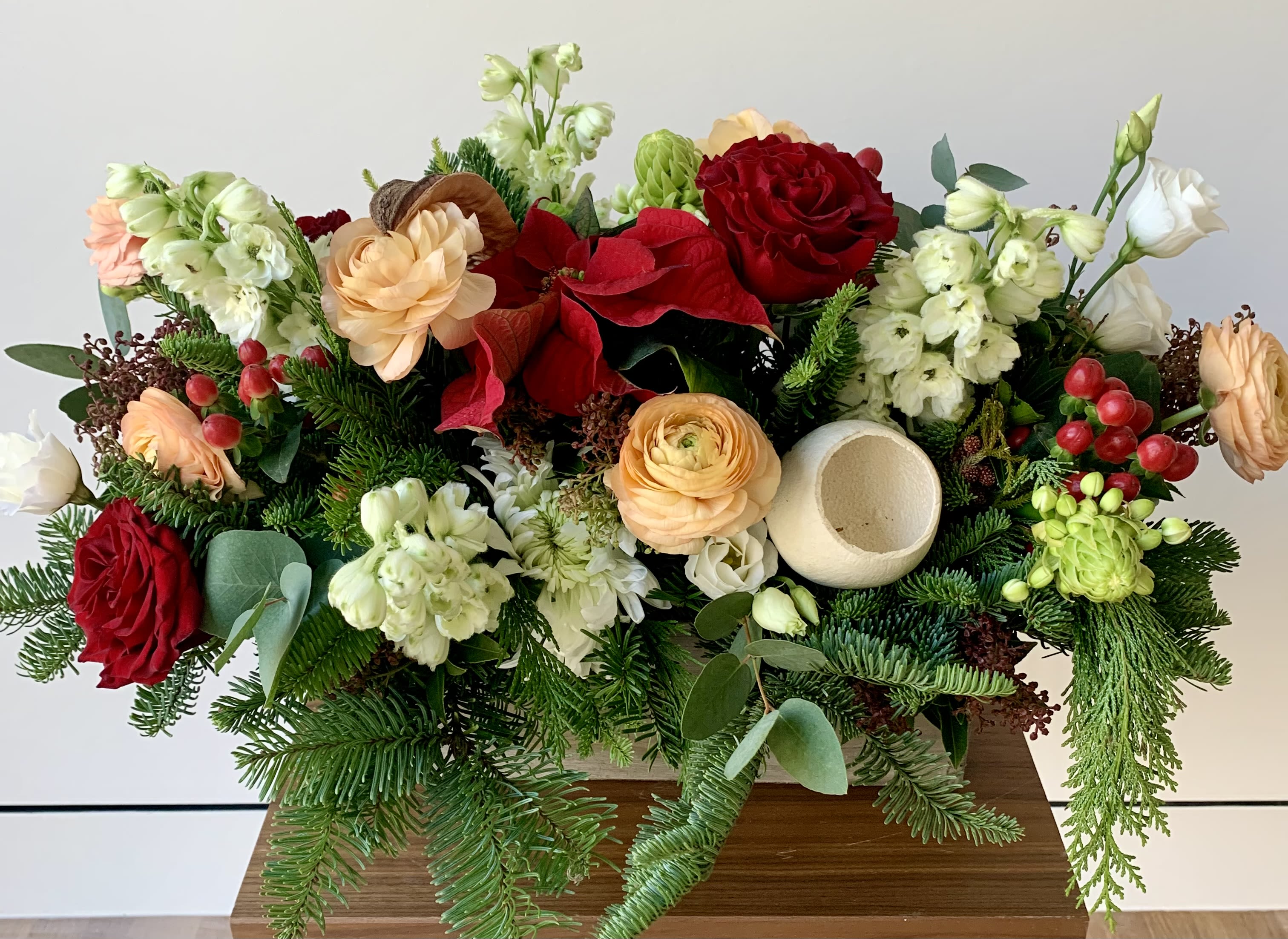 Floral Ornament - Lovely holiday flower arrangement in a rustic wooden box 12x4x4 in softer festive color tones with a pop of red. A mix of wintergreens, berries, roses, ranunculus, lisianthus, delphinium, white star and additional flower fillers. 