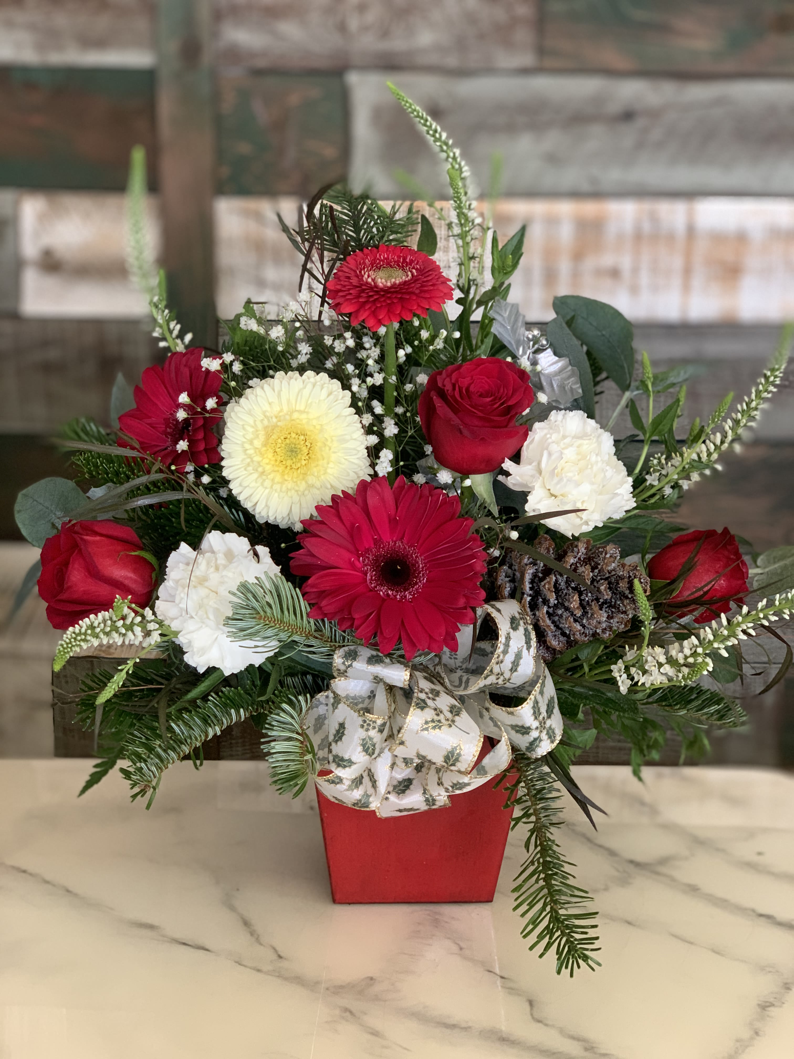 Holly and Bright designers choice - Step into the festive season with Holly and Bright. This darling arrangement captures the essence of Christmas joy, boasting vibrant red roses and carnations, gerberas, veronica  and lush evergreens, all coming together to evoke the cozy warmth of the holidays. Nestled playfully among the blooms is a pinecone for a rustic element, reminiscent of a winter walk in the woods, while the varied greenery provides a fresh, full backdrop that makes the red and white colors truly pop. Presented in leakproof red cube this arrangement is a nod to the traditional yuletide palette, making it a perfect centerpiece for holiday gatherings or a cheerful addition to any room.  