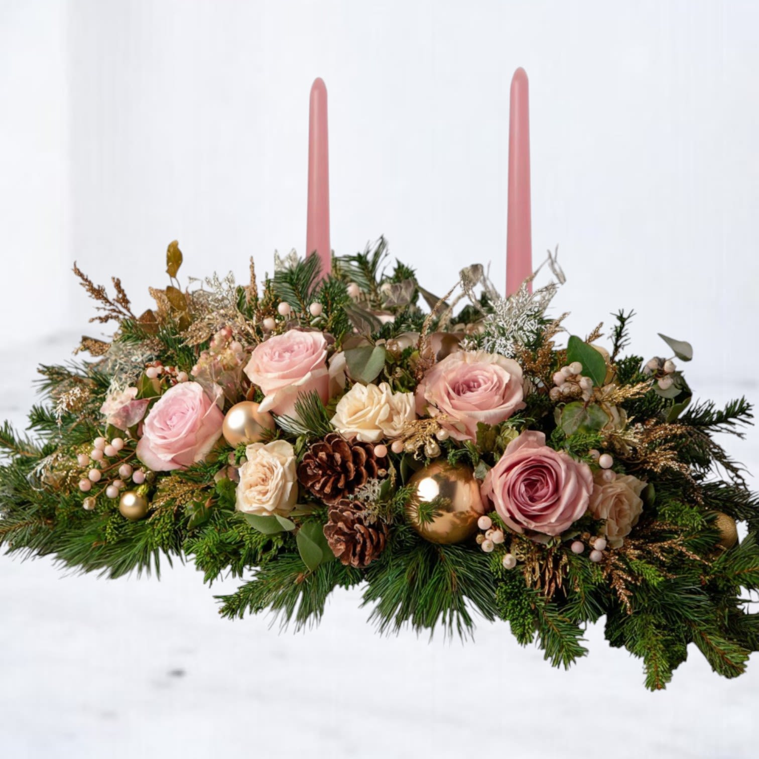 Blush Solstice Centerpiece - The Blush Solstice Centerpiece blends romantic winter tones with soft seasonal elegance. Designed with blush roses, creamy spray roses, winter greenery, pinecones, champagne ornaments, and delicate holiday textures, this arrangement glows with warmth and sophistication. Two blush taper candles add a gentle, welcoming light — perfect for holiday gatherings, intimate dinners, or festive winter décor.  Handcrafted by Whiskey Wallflower, this design brings a fresh, modern twist to traditional holiday centerpieces, combining soft florals with luxe accents for a beautifully balanced winter statement piece.  Available for same-day holiday and Christmas delivery in Magnolia, The Woodlands, Conroe, Tomball, and surrounding Houston communities.  Substitution Disclaimer: Seasonal flowers or accents may be replaced with premium alternatives of equal or greater value while maintaining the overall look and palette.