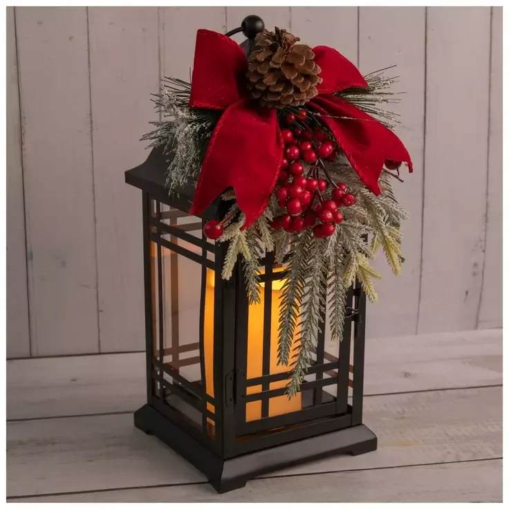 Winterberry Ribbon Glow Lantern - Bring festive elegance to your holiday décor with this stunning black metal lantern, topped with a lush red velvet bow, frosted greenery, bright berries, and a rustic pinecone. The warm LED candle inside casts a cozy glow, making it a perfect accent for entryways, mantels, or holiday centerpieces. 13.75&quot; H x 8.5&quot; W x 9.5&quot; D