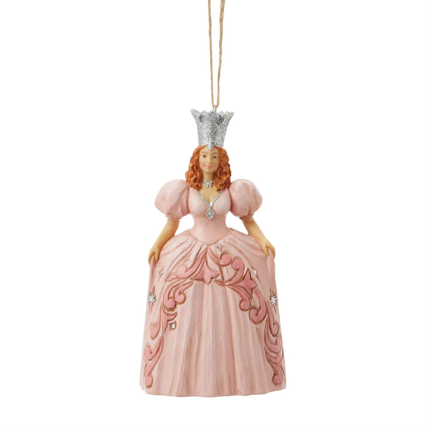 Glinda Ornament - Celebrate the enchantment of the season with this officially licensed Wizard of Oz Glinda the Good Witch ornament. Radiant in her iconic blush-pink gown, Glinda brings warmth, wonder, and a touch of cinematic nostalgia to your holiday display. Her sculpted gown is adorned with swirling filigree and sparkling star accents, all crowned by her signature silver tiara that catches the light beautifully.  As part of the Four Realms of Christmas, Glinda embodies the Realm of Magic—guiding the season with grace, protection, and charm. Whether adorning your tree or adding a whimsical centerpiece to your holiday décor, she infuses every moment with the gentle glow and timeless elegance that fans have cherished for generations. Crafted with hand-painted detail and suspended by a classic twine hanger, this piece is a keepsake meant to be treasured year after year.