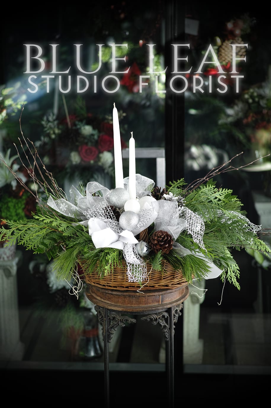Snow and Cedar - Lush, fragrant evergreens line a festive wooden basket filled with white and silver tufts of ribbon, pinecones, and white taper candles, creating the perfect holiday centerpiece. 