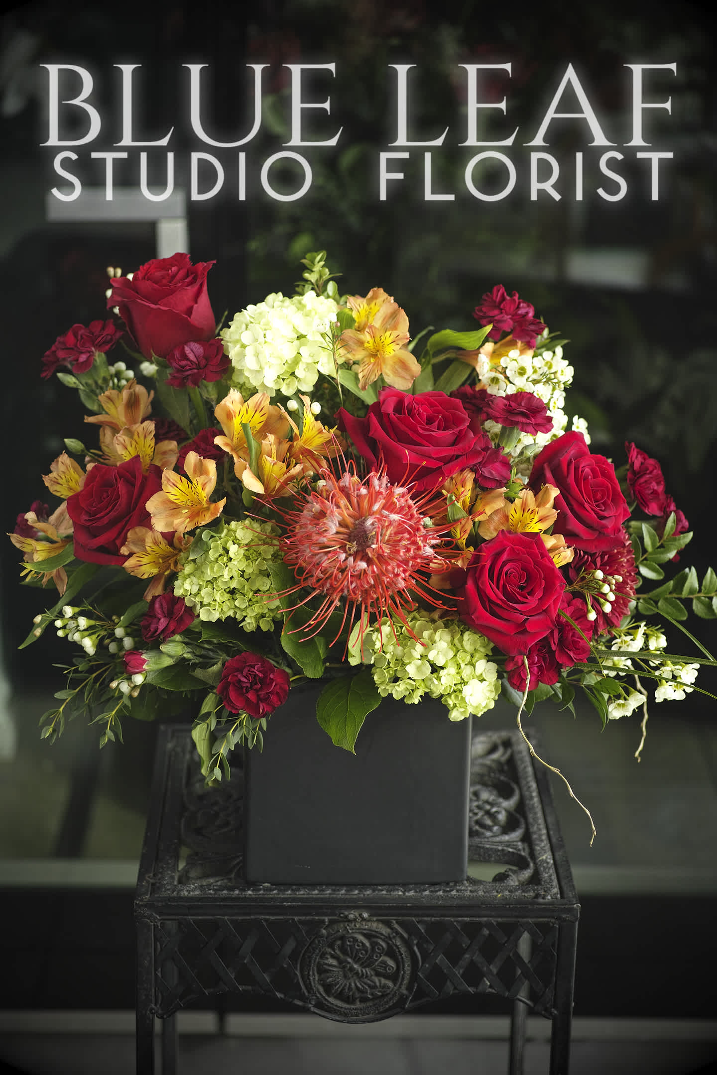 Warm Regards - Sophisticated floral arrangement  of red roses and a mixture of warm tone flowers. Designed one sided in a ceramic cube. All around is available in the Premium selection.