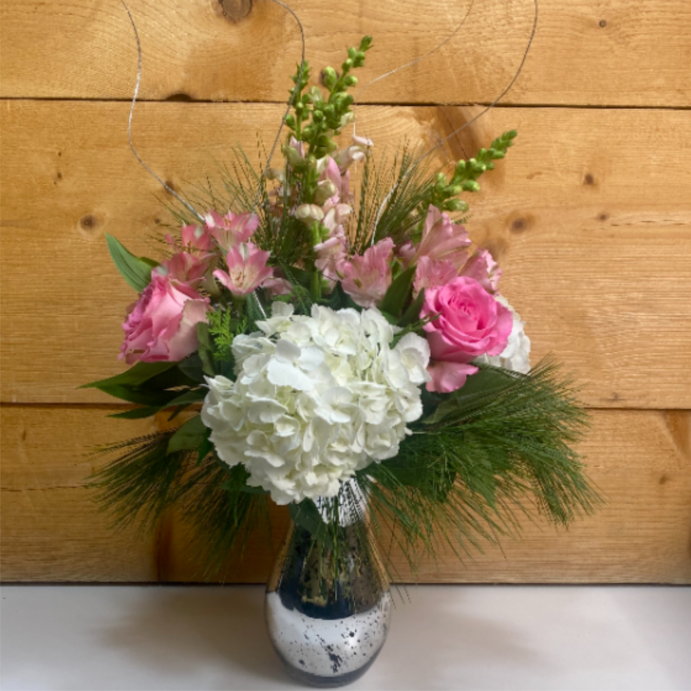 Pretty in Pink Holiday Bouquet - The Pretty in Pink Holiday Bouquet is a charming arrangement that combines a wintery touch with blushing pink flowers including roses, snapdragons, and alstroemerias. Artfully arranged in a silver mercury glass vase with silver ting for decoration, this arrangement is perfect for any joyous occasion in the winter, not just for Christmas!  Dimensions approx. L 16&quot;x H 29&quot; (with ting!)
