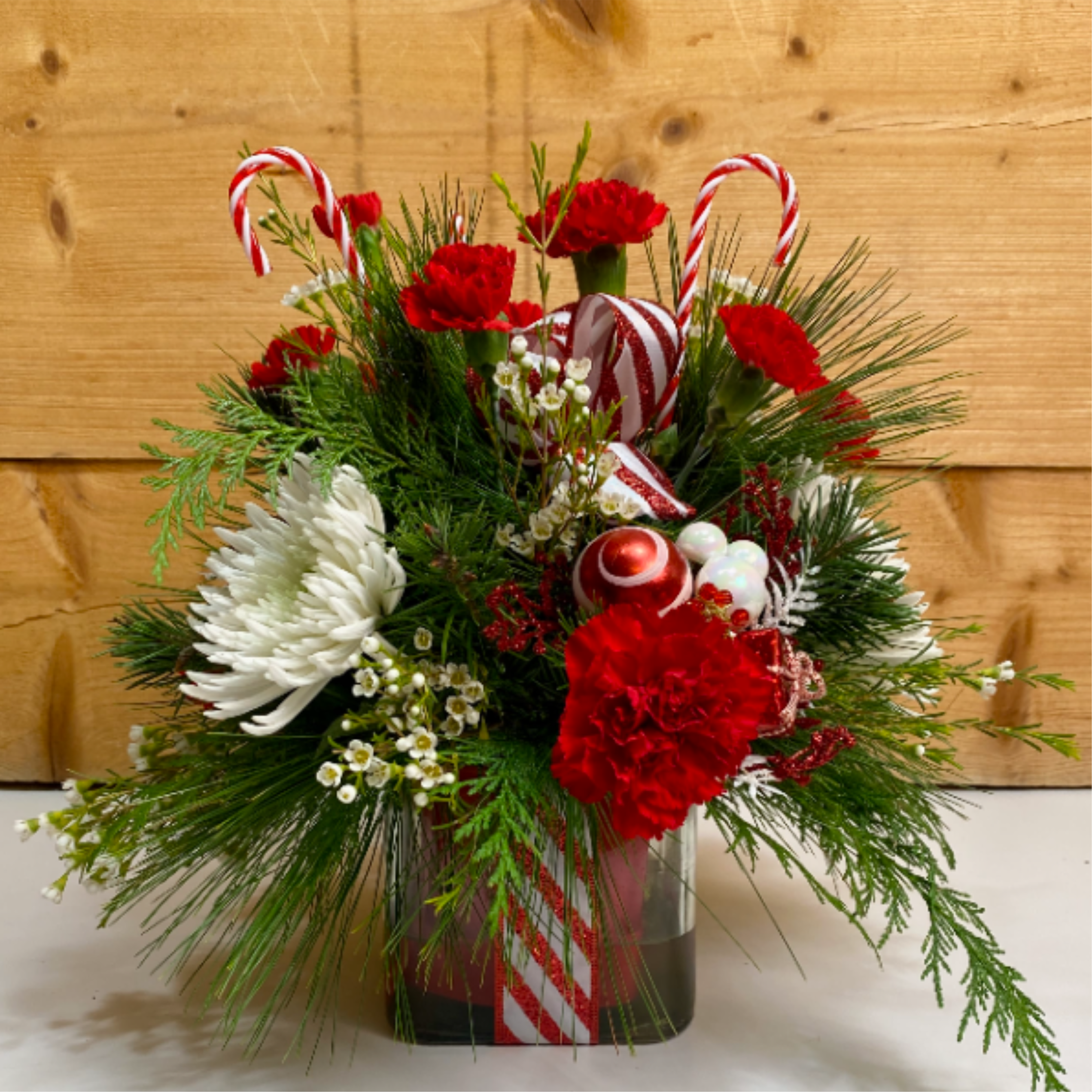 Candy Cane Lane - Candy Cane Lane is a fun arrangement that brings peppermint perfection into your home! Presented in a glass cube with candy cane ribbon and a glittery candy cane bow, this arrangement features red carnations and white mums and waxflower, along with decorative candy cane themed picks for a delightful design that's sure to bring joy this festive season!  Dimensions approx: L 14&quot; x H 15&quot;