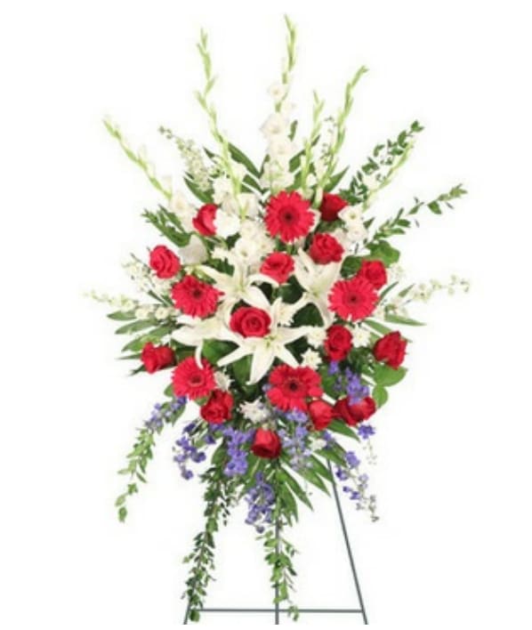 Brave Salute Easel - This stunning sympathy tribute is gracefully presented on a 54-inch easel, capturing both elegance and strength in its design. At its heart is a large wet floral foam cage that supports a lush array of foliage, including myrtle, Teepee Emerald, and salal, providing a rich green backdrop that enhances the beauty of the blooms. Three pristine white lilies signify purity and devotion, while five majestic white gladiolus convey strength of character. In vibrant contrast, five red gerberas and twelve striking red roses symbolize love and remembrance, infusing the arrangement with passion and warmth. Complementing this heartfelt display are three delicate stems of white larkspur, which suggest an open heart and joy, alongside three stems of fragrant white stock that express lasting beauty. The addition of seven vibrant blue delphiniums brings a calming touch, while three white cushion poms add a sense of softness and serenity. Together, this arrangement stands as a touching homage to a cherished life, providing comfort and solace to all who view it.