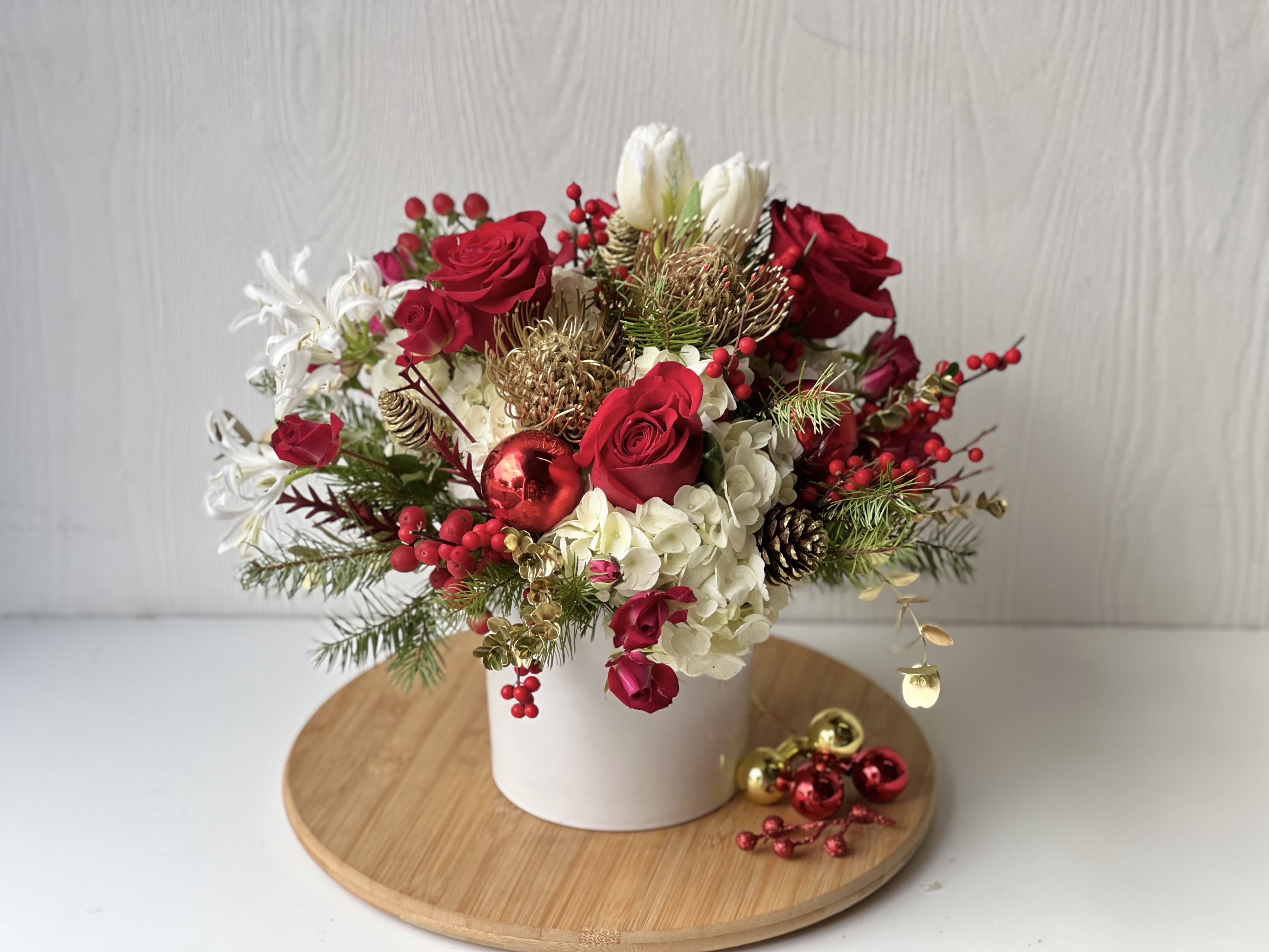 Festive Luxe Blooms - Celebrate the season with the Festive Lux Blooms, a stunning holiday arrangement that embodies the spirit of Christmas. This luxurious floral design features lush white hydrangeas, vibrant red roses, and elegant white tulips, accented by striking pincushion proteas. Nestled among fragrant Christmas greenery, the arrangement is adorned with chic holiday accents in gold, adding a touch of festive opulence. Perfectly presented in a modern white ceramic vase, this bouquet radiates sophistication and holiday cheer.  Whether as a show-stopping centerpiece or a heartfelt gift, the Golden Noel Elegance is crafted to bring joy, elegance, and seasonal beauty to any space. Order now for a memorable holiday statement!