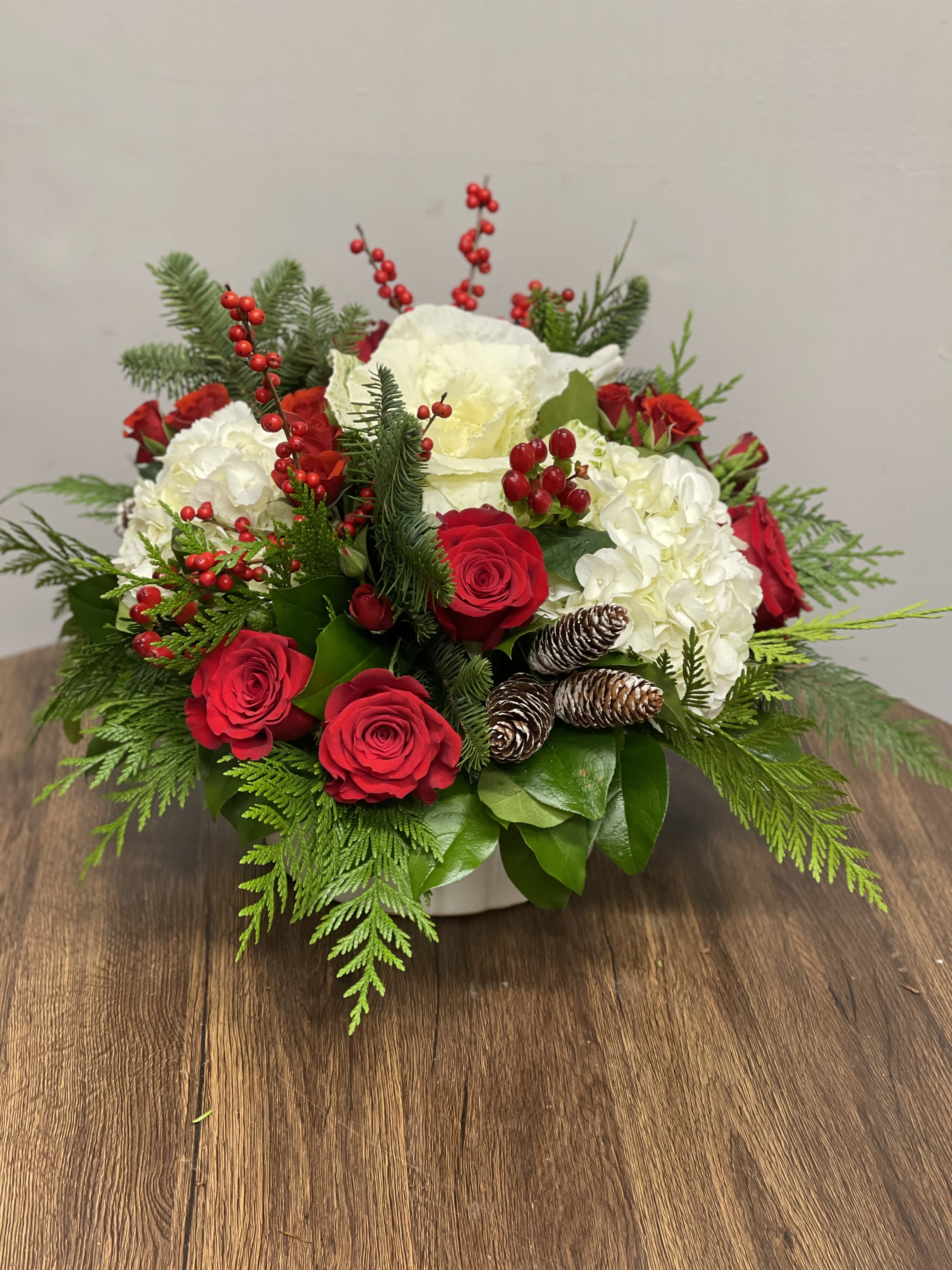Classic Holiday - Beautiful combination of white Hydrangeas, red Roses, white Kale and other seasonal flowers with Winter greens and pine cones in a round ceramic container.
