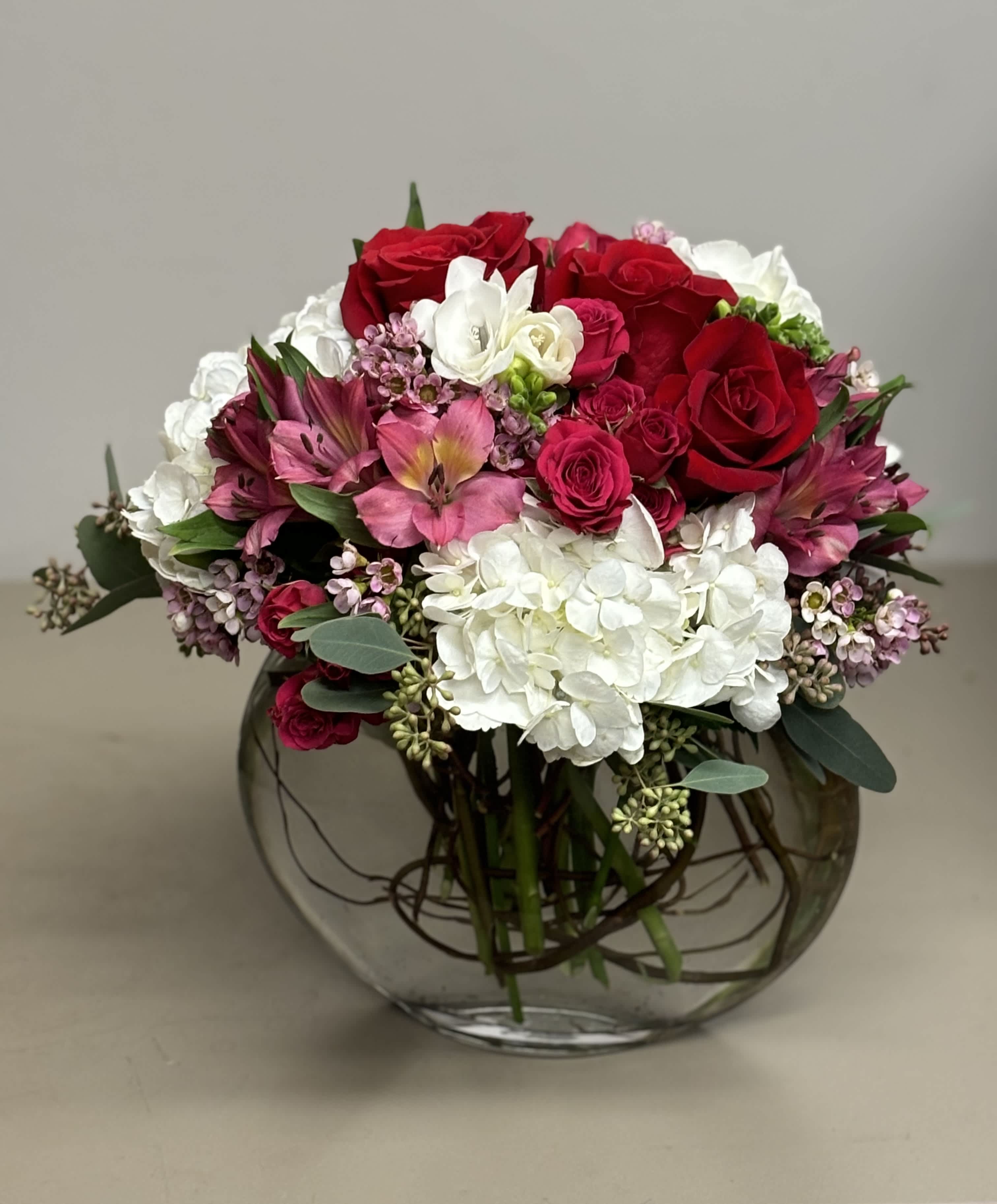 Cupid - Beautiful combination of white Hydrangeas, red Roses, hot pink spray Roses and other seasonal flowers in an oval shaped glass vase.