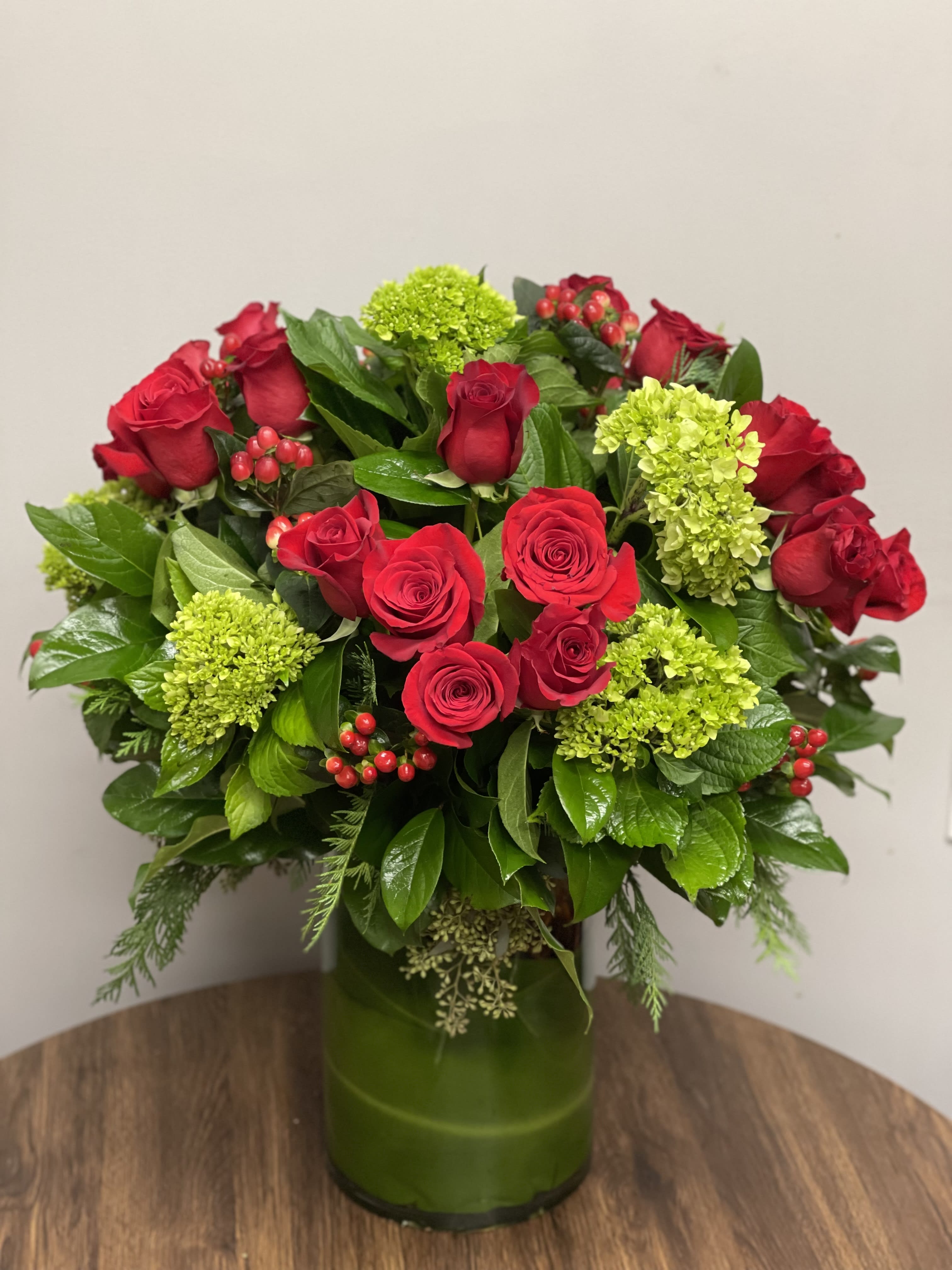 Holiday Cheer - A beautiful holiday arrangement with  2 dozen red Roses, Green Hydrangeas, Red Hypericum berries and other seasonal greens in a glass cylinder vase.