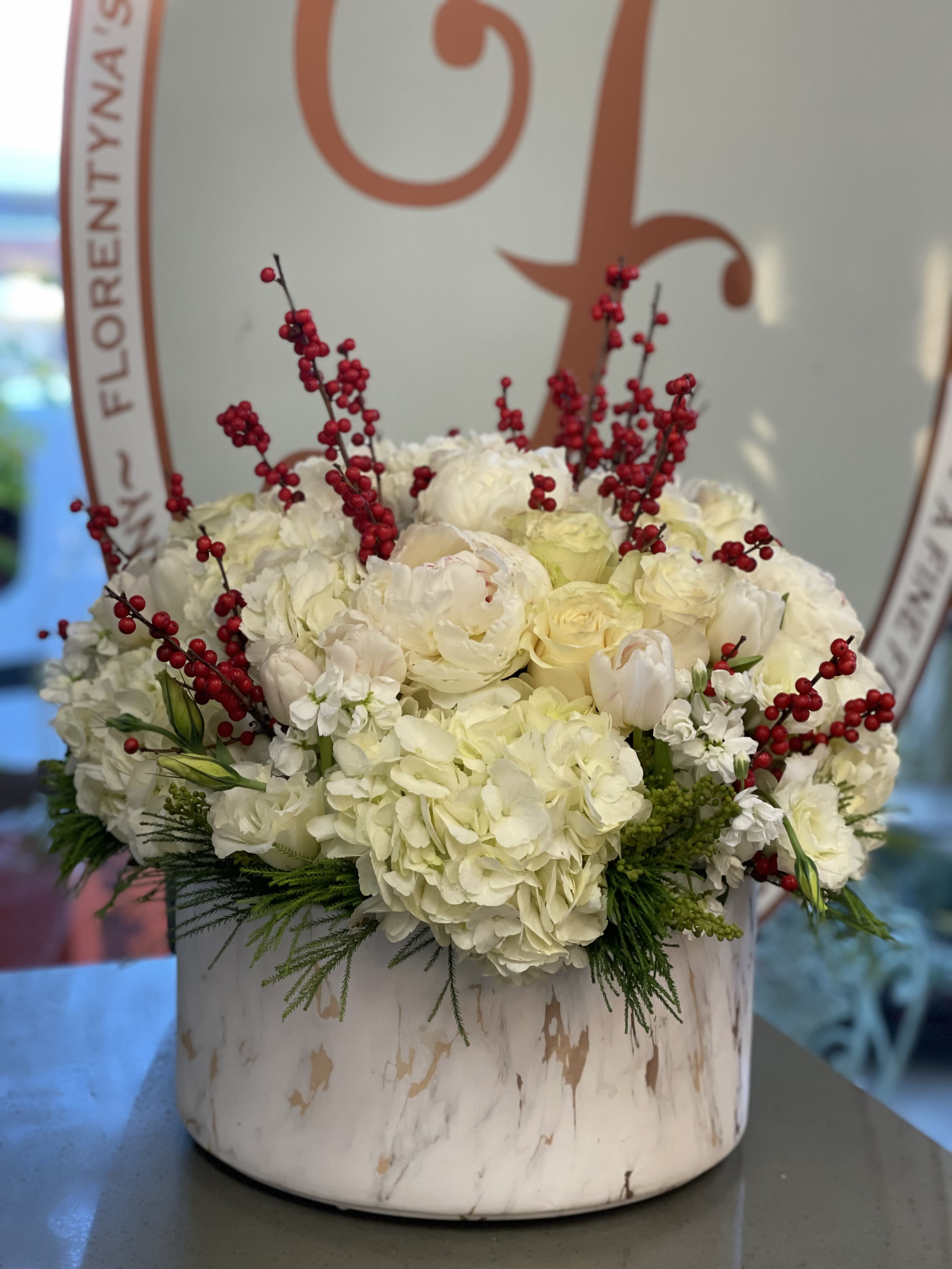 Navidad - Beautiful combination of white Hydrangeas, white roses, white Peonies, red berries  and other seasonal flowers and Winter greens in white round ceramic container. ***containers might change due to availability***
