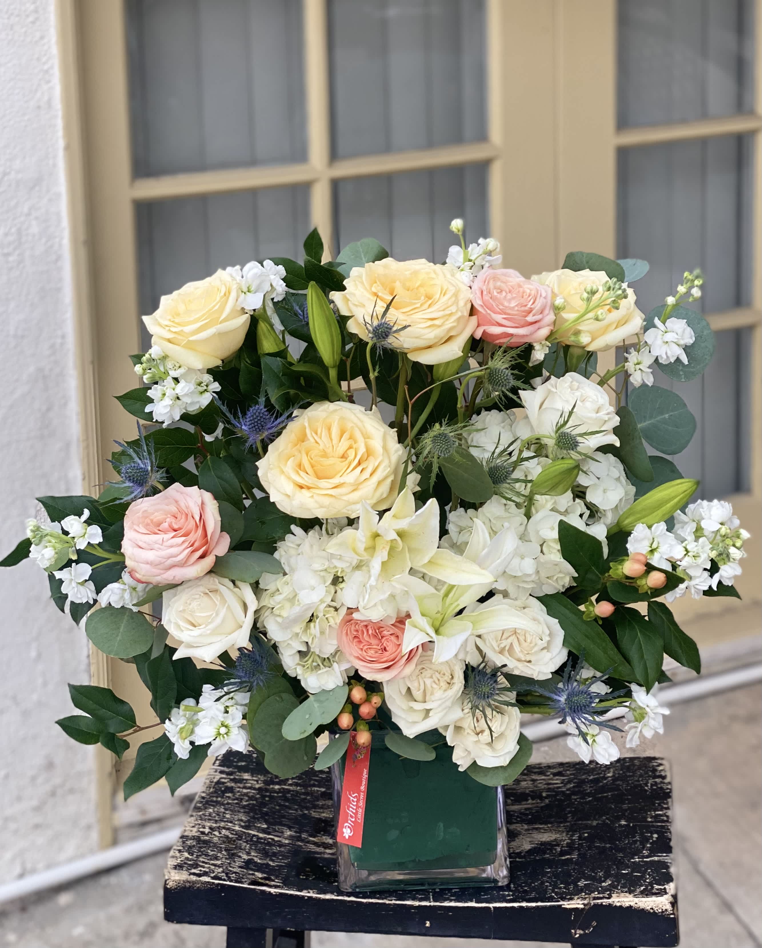 Garden Peach Roses Candle Light Roses Hydrangea Modern Garden Cube Vase Arrangement  - Arrangement Measures approximately 22"H  Send a  a bit of modern style garden arrangement! It includes white hydrangea premium candle light  roses, garden peach roses, casa blanca lilies, and blue eryngium accented with fresh greenery and delivered in a contemporary glass cube.  Perfect gift to celebrate Mother's Day, a birthday, to decorate the stage of an event, dress up your podium or memorial services or just because!  