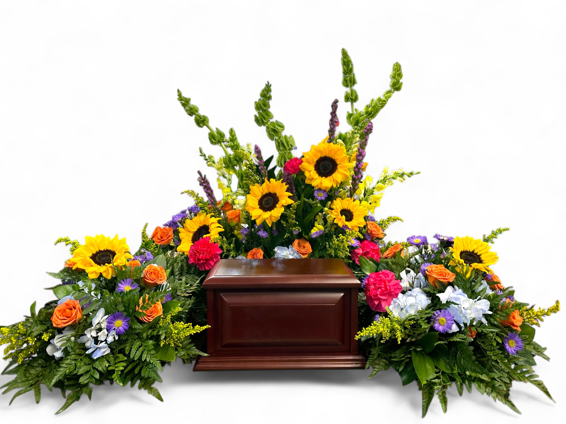 Vibrant Life Urn Tribute - This heartfelt tribute arrangement features a vibrant display of sunflowers, roses, hydrangeas, and other colorful blooms, thoughtfully arranged around a polished wooden urn. The bright yellow sunflowers symbolize admiration and gratitude, while the orange and purple flowers add warmth and serenity. Lush greenery frames the arrangement, creating a comforting and respectful tribute that celebrates a life filled with beauty and joy. Perfect for honoring and remembering loved ones with dignity and love.