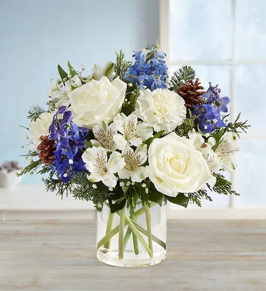  Winter Wishes Bouquet - Inspired by winter’s cool white &amp; blue landscape, our charming arrangement captures every wish you want to express to those who mean the most. Hand-designed inside a clear cylinder vase, this is one gift they’ll be talking about long after the season’s over. •All-around arrangement with white roses, carnations and Peruvian lilies (alstroemeria), blue delphinium; baby’s breath; seeded eucalyptus and assorted greenery and pinecones •Peruvian lilies may arrive in bud form and will open to full beauty in 2-3 days