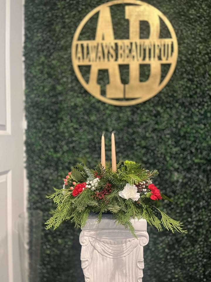 Christmas Centerpiece Decorated with Candles - This Christmas centerpiece is a lush and elegant arrangement crafted entirely from vibrant Christmas greens, beautifully adorned with festive flowers. A rich mix of fragrant evergreen branches, including pine, fir, and spruce, creates a full, textured base, evoking the spirit of a winter forest. Sprigs of holly with their glossy, spiked leaves add a touch of festive contrast, while the deep green of boxwood enhances the centerpiece’s refined look. Subtle sprigs of cedar lend a soft, feathery texture, while seasonal blooms, such as red poinsettias, white lilies, and delicate roses, bring vibrant color and a touch of holiday cheer. While candles nestled within the greens provide a gentle, flickering light, creating a cozy, enchanting atmosphere. This centerpiece brings a peaceful, organic touch to your table, capturing the essence of Christmas with its evergreen beauty and warm candlelight. This centerpiece combines the beauty of fresh greens and the charm of seasonal flowers, creating a stunning and joyful addition to your holiday table.