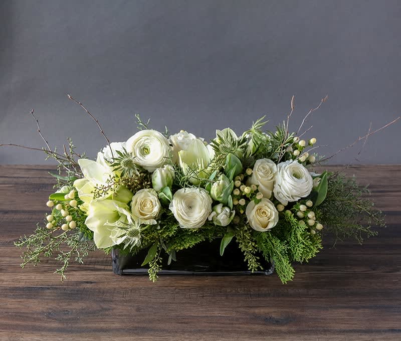 Snow White Centerpiece  - This year, make the holidays bright with a showstopping centerpiece. Snowy amaryllis blossoms, radiant ranunculus, and immaculate white tulips emerge from a fresh backdrop of evergreen textures and silvery thistle. Crisp ivory roses, helleborus ‘winterbells,’ and towering birch branches complete the tasteful design. Artfully arranged in a long, leaf-lined vase.   You deserve the best. If an item is unavailable, we’ll sub in products of equal or greater value.