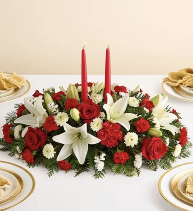 Winter Harmony Centerpiece - A serene, festive centerpiece that feels like the quiet magic of a holiday evening. Two slender red taper candles rise from a lush bed of seasonal evergreens, casting a warm glow across a tapestry of blooms. Pure white lilies open like winter stars, their soft petals adding elegance and light, while rich red roses and cheerful red carnations bring depth, romance, and classic holiday warmth. Accents of white cushion mums weave through the arrangement like gentle snowflakes, adding texture and balance. Designed low and elongated for the holiday table, this centerpiece creates an atmosphere of celebration, togetherness, and calm winter beauty. A perfect statement for Christmas dinners, winter gatherings, or any festive moment where you want the room to feel warm, joyful, and full of light.