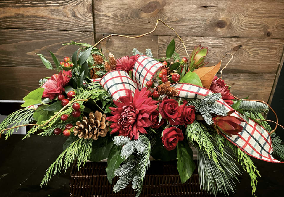 Plaid &amp; Pine Holiday Long Centerpiece - This lush, elongated holiday centerpiece is full of classic Christmas charm. Rich red blooms—roses, mums, and alstroemeria—are nestled among mixed evergreens, pinecones, red berries, and frosted foliage for a natural winter woodland feel. The bold plaid ribbon adds a cozy, traditional touch, while curly willow brings movement and organic texture. Designed long and low, it’s ideal for a dining table or buffet, creating a warm, festive focal point for holiday gatherings.