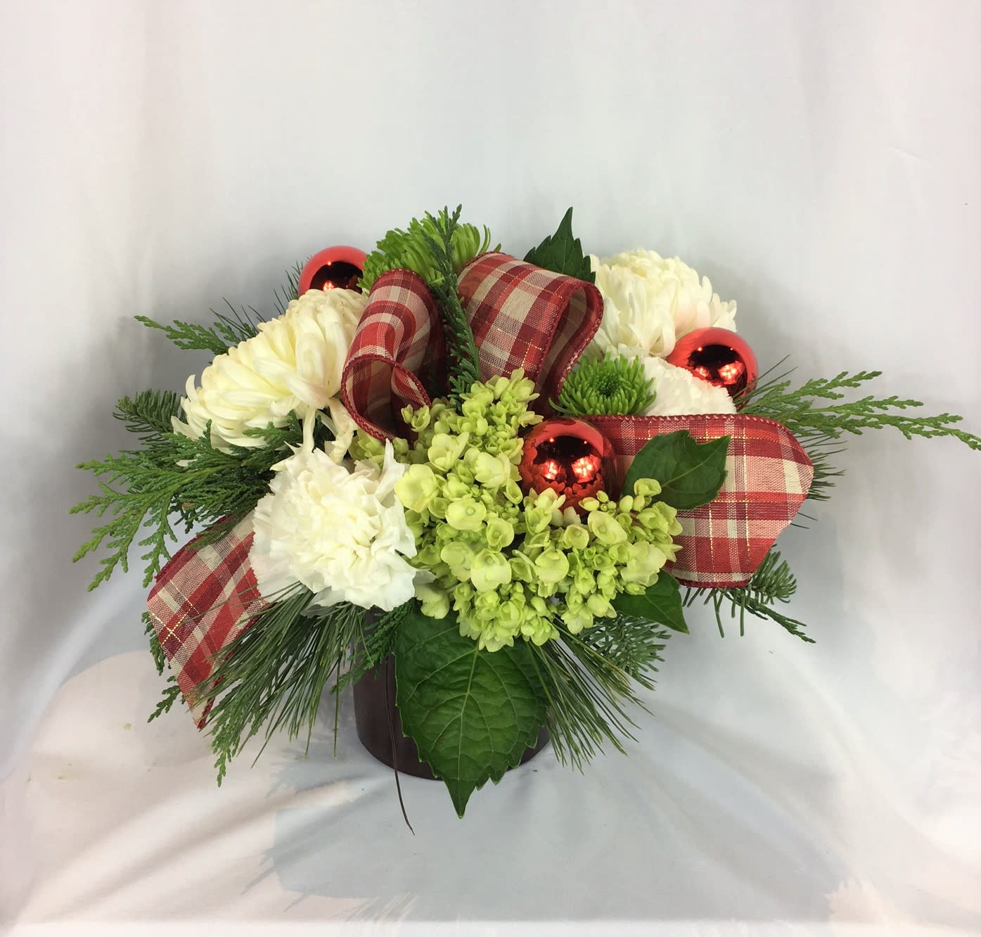 Frosted Woodland Bouquet - This arrangement blends crisp winter whites with fresh, natural greens for a clean and festive look. White hydrangea and mums create a soft, snowy feel, while green mini mums and seasonal foliage add depth and texture. The plaid ribbon introduces a cozy, rustic holiday touch, complemented by shiny red ornament accents for a cheerful pop of color. Designed in a compact, rounded style, it’s perfect for holiday gifting or seasonal home décor.