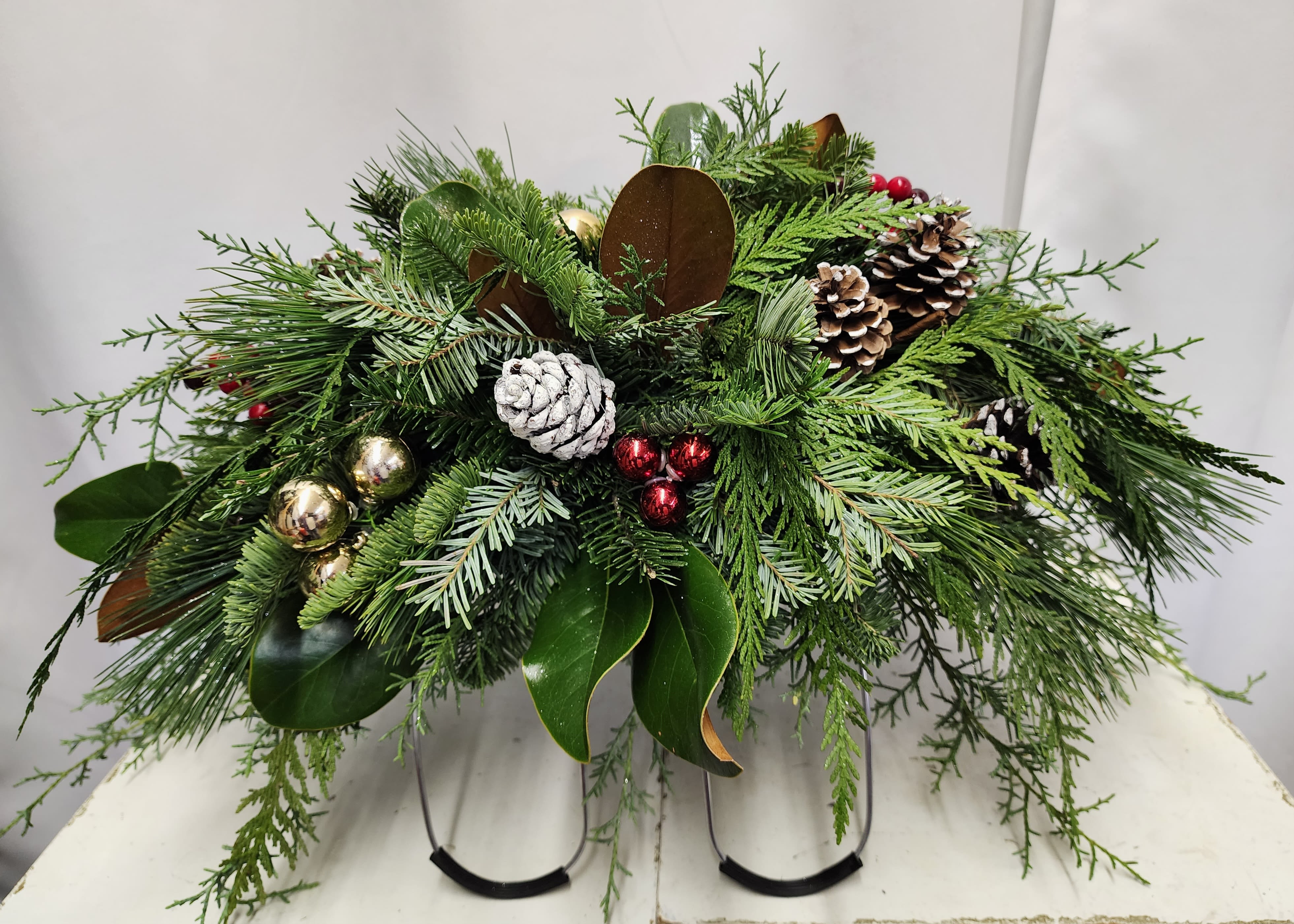 Holiday Headstone Cover - Bring festive warmth and remembrance to your loved one's resting place with our Holiday Headstone Cover. Crafted with fresh holiday greens, vibrant berries, decorative ornaments, and natural pinecones. This cover adds a touch of seasonal beauty to any cemetery headstone. Each piece is arranged on a sturdy wire frame featuring protective rubber feet that can be securely tightened to fit any headstone size. Custom options are available—add fresh flowers or ribbons for an extra special touch. Please note that the finished product will closely resemble the sample shown based on greens available. Limited quantities and requires 24 hours notice.  In store pick up only. 