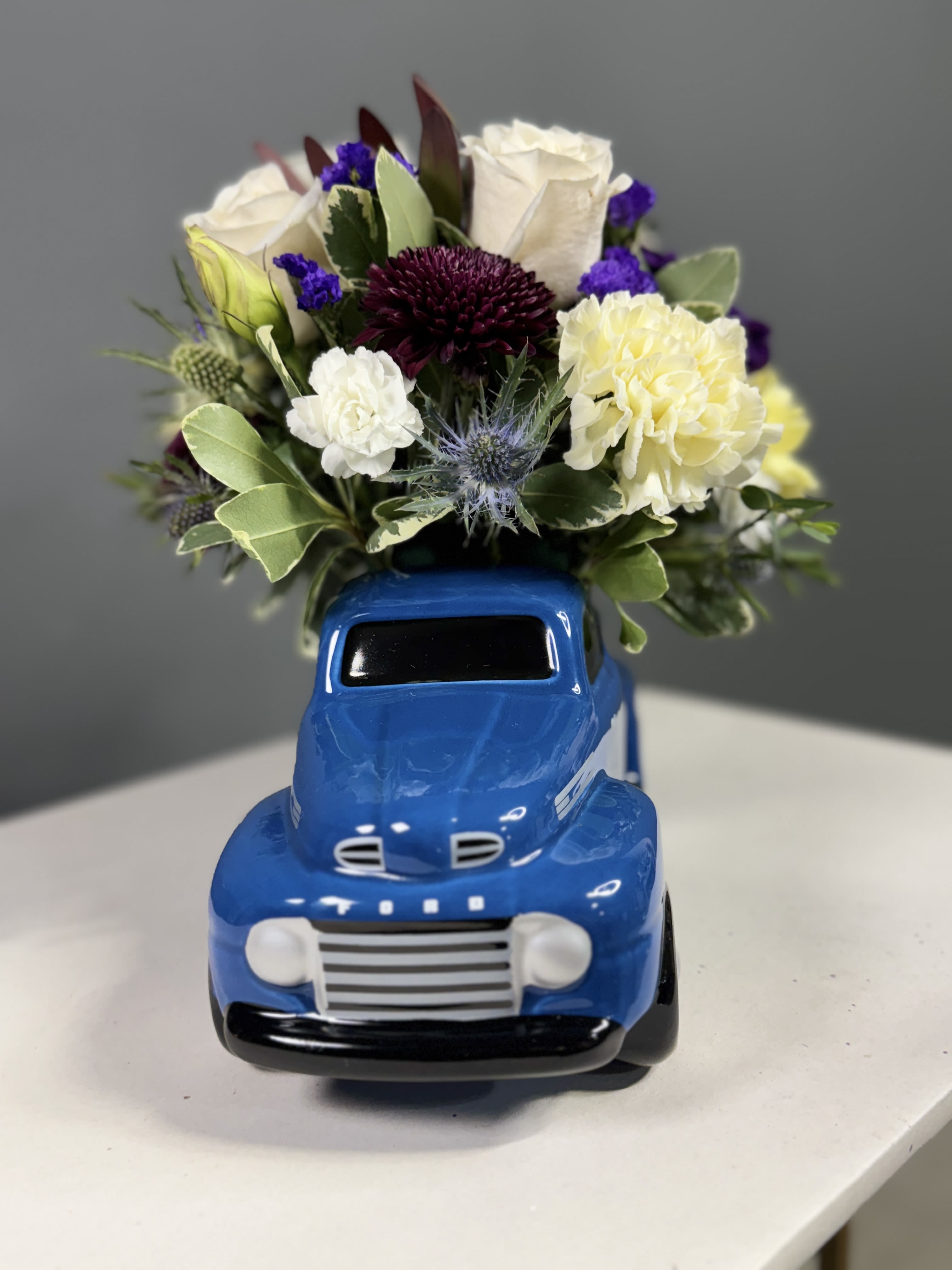 Vintage Ford Pickup -  "Vintage Ford Pickup" floral creation, a charming and whimsical ensemble that blends rustic charm with floral elegance. Nestled within the confines of a blue '48 Ford pickup ceramic keepsake, this arrangement features a delightful mix of white roses, white, purple, burgundy, and creme flowers, accented with a touch of blue blooms. The Vintage Ford Pickup arrangement embodies a sense of nostalgia and countryside allure, making it a delightful centerpiece that evokes memories of simpler times and adds a touch of rustic sophistication to any space.