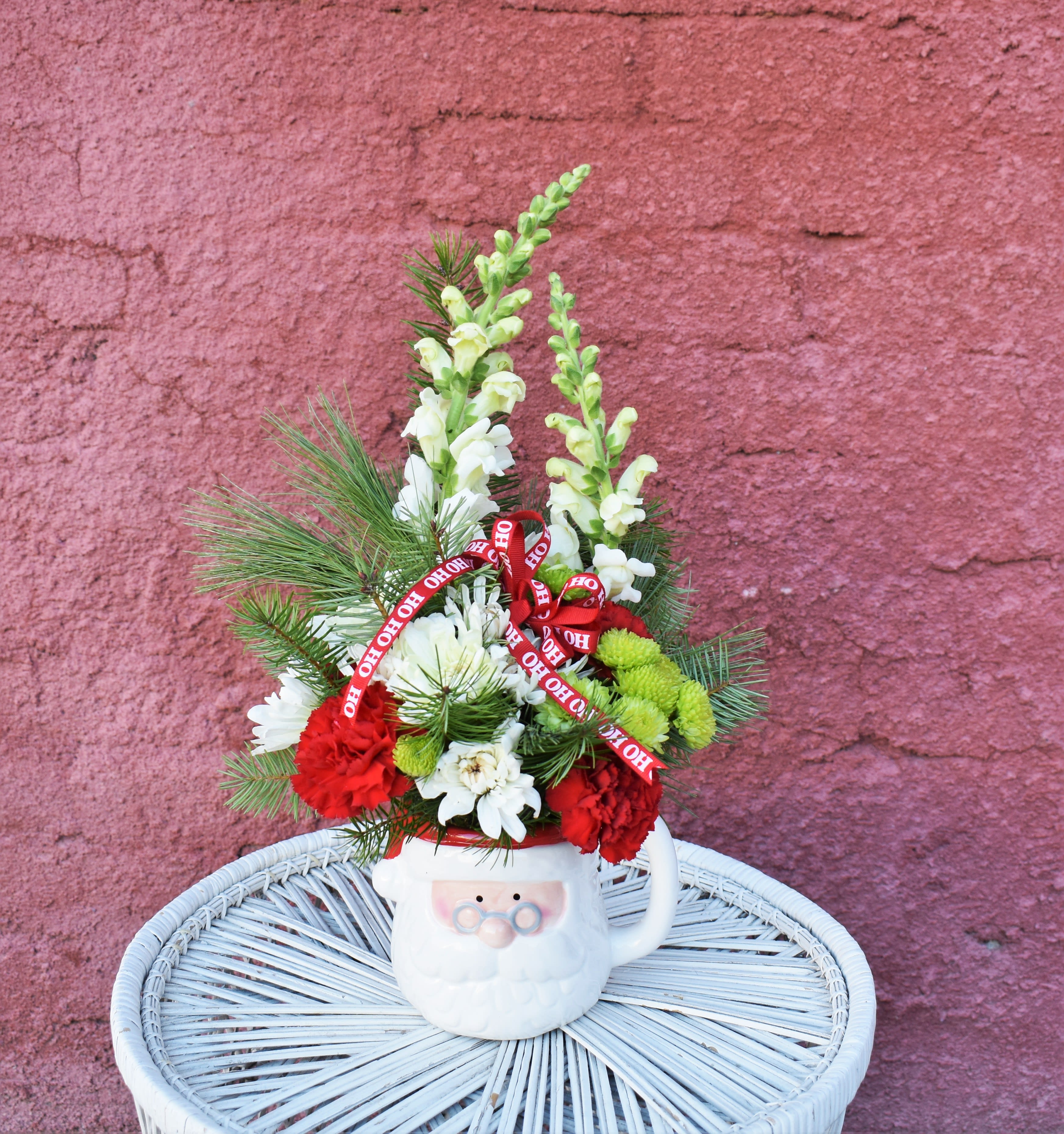 Santa Baby - This cute mug means that Santa Claus must be coming to town! White snapdragons, red carnations, white mums, and green button mums are designed in an adorable Santa mug and accented with a Christmas bow...a fun way to celebrate the season! 