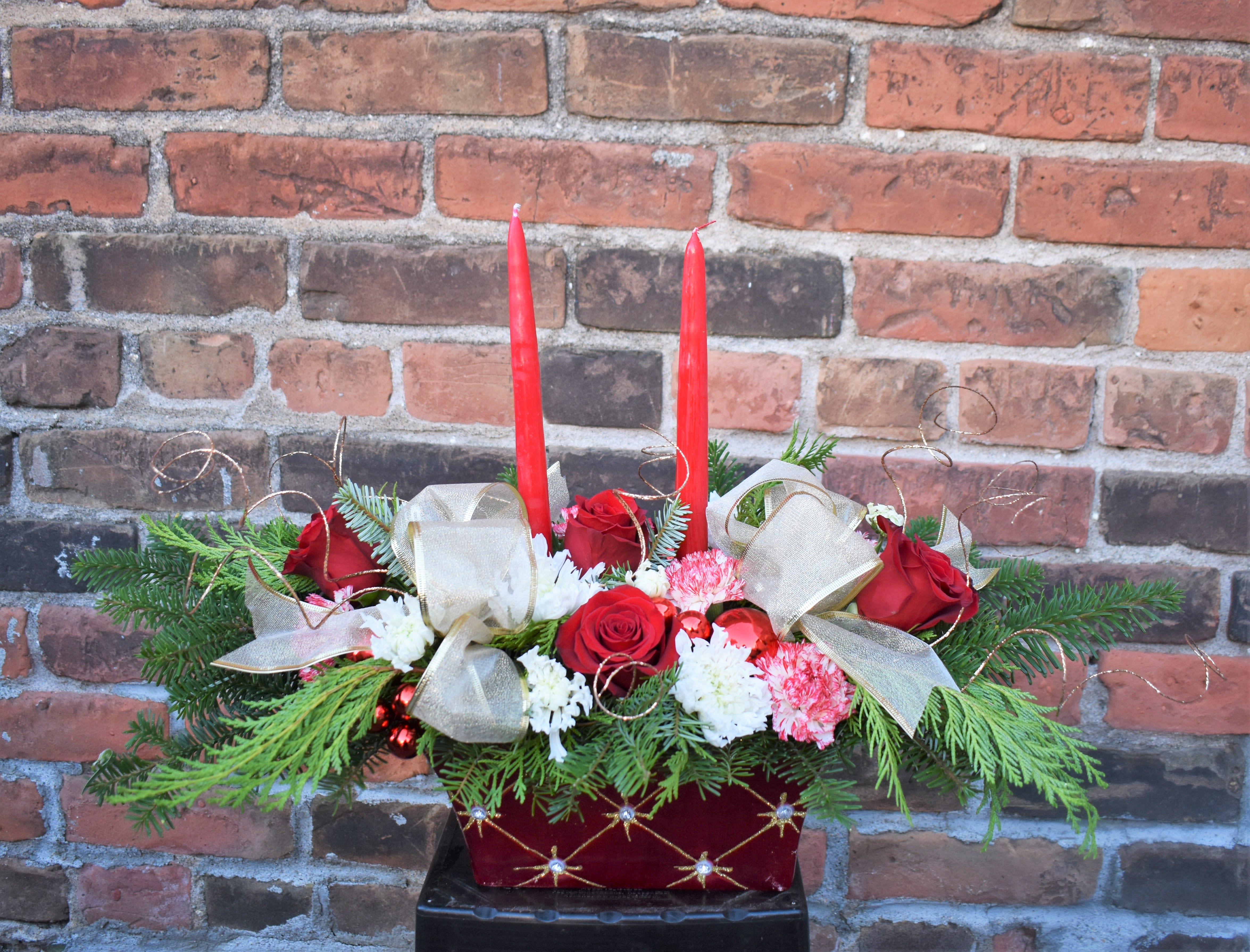 Joy to the World - This centerpiece is full of Christmas joy! Red roses, peppermint carnations, white mums, and fresh pine are designed in a red ceramic container and accented with gold bulbs, ribbon, and ting ting. The red ceramic container is accented by sparkling rhinestones. Two candles complete the design and create a fun centerpiece for any Christmas gathering! 