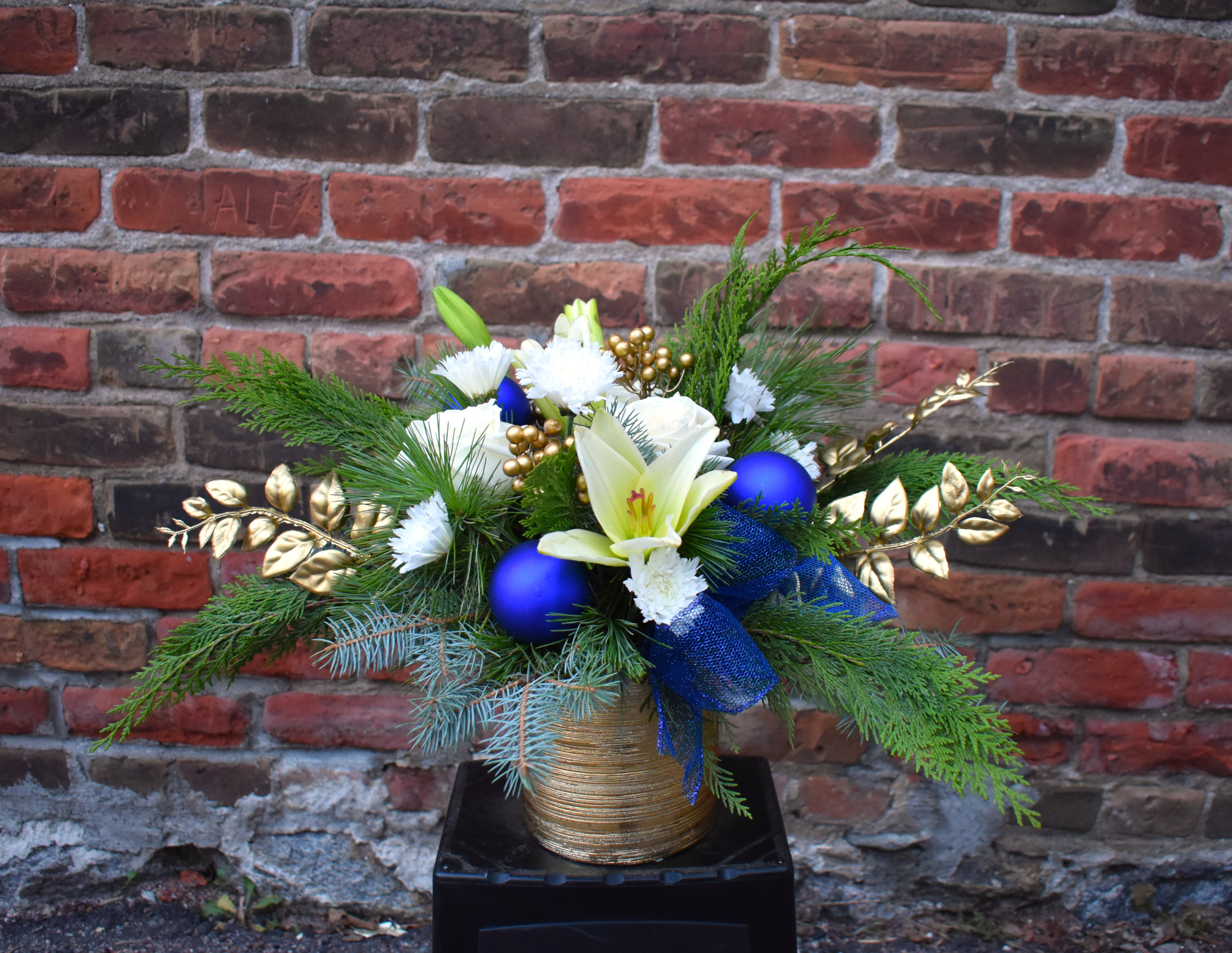 Noel - Regal blues and bright whites make this an eye-catching design! White lilies, cushion mums and roses are designed in a gold ceramic container and accented with fresh pine, gold branches, and blue ornaments...A peaceful and gorgeous way to make a statement this Christmas! 