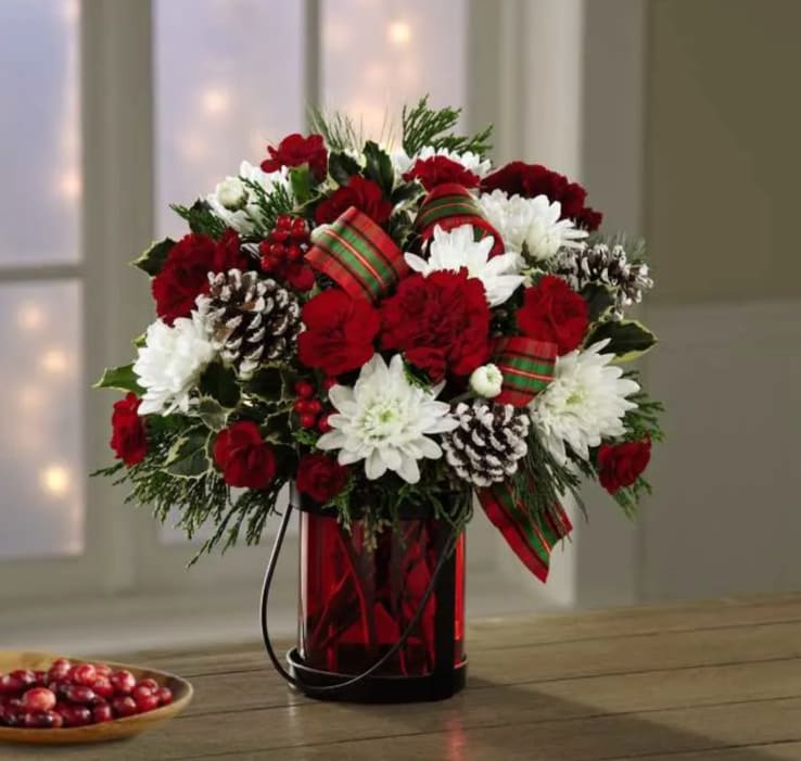 Holiday Wishes - Evoking the warmth and joy of a cozy Christmas, this bouquet delivers your warmest season's greetings. Composed of white cushion pompons, red carnations, Christmas greens with holly berries, and &quot;frosted&quot; pinecones. This arrangement is accented with bright tartan bows in a ruby-hued glass vase.