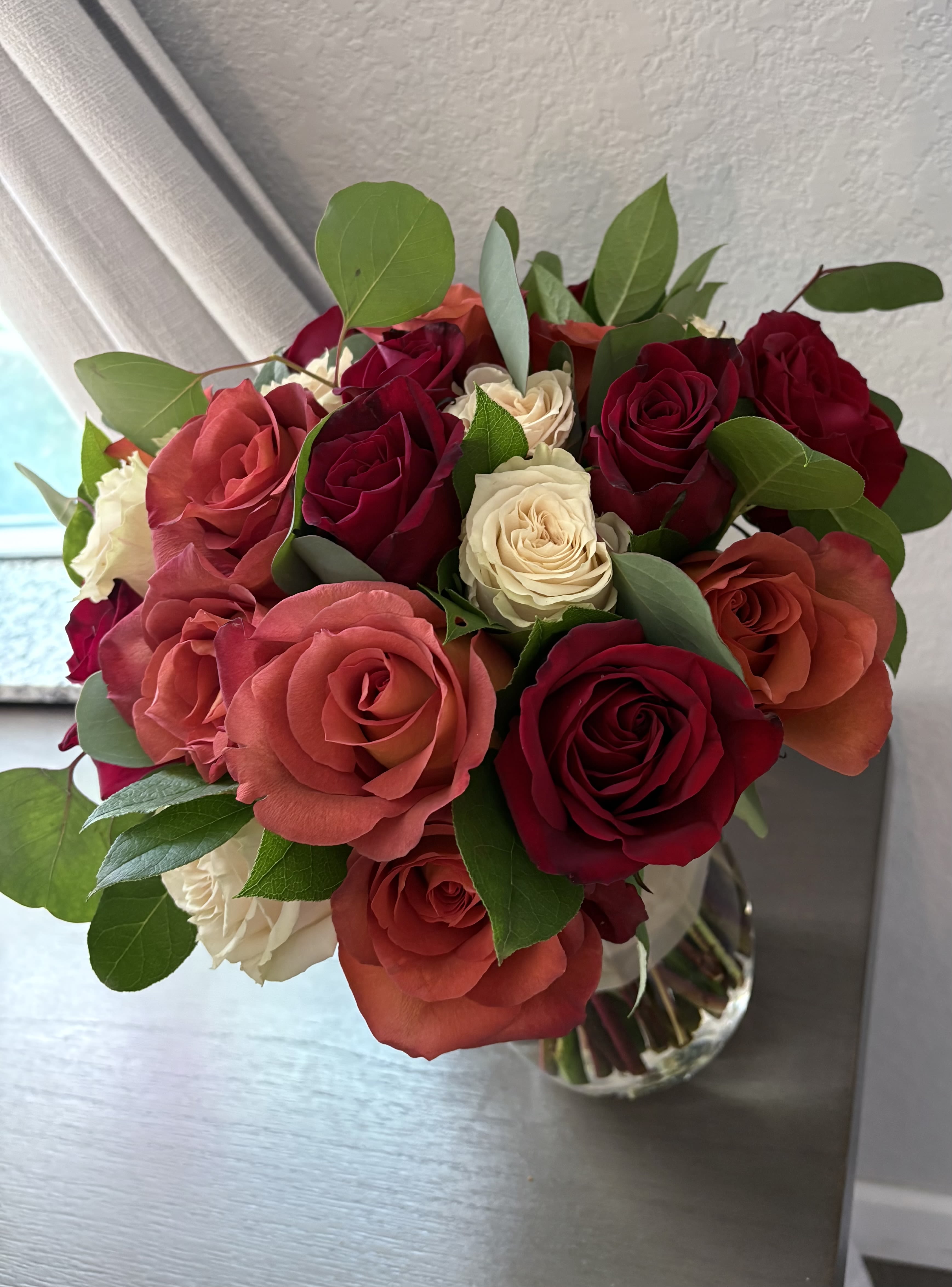 Crimson Dawn - "Crimson Dawn" is an enchanting floral arrangement that embodies the vitality of a new day. This delightful bouquet features a vibrant array of roses in deep red, lively orange, and subtle cream hues, each blossom exuding a lush and velvety texture. The clear glass vase enhances the bouquet's natural elegance, while the surrounding green leaves add refreshing contrast. Positioned by a softly lit window, "Crimson Dawn" infuses any space with warmth and charm, reminiscent of a serene morning bathed in gentle sunlight.