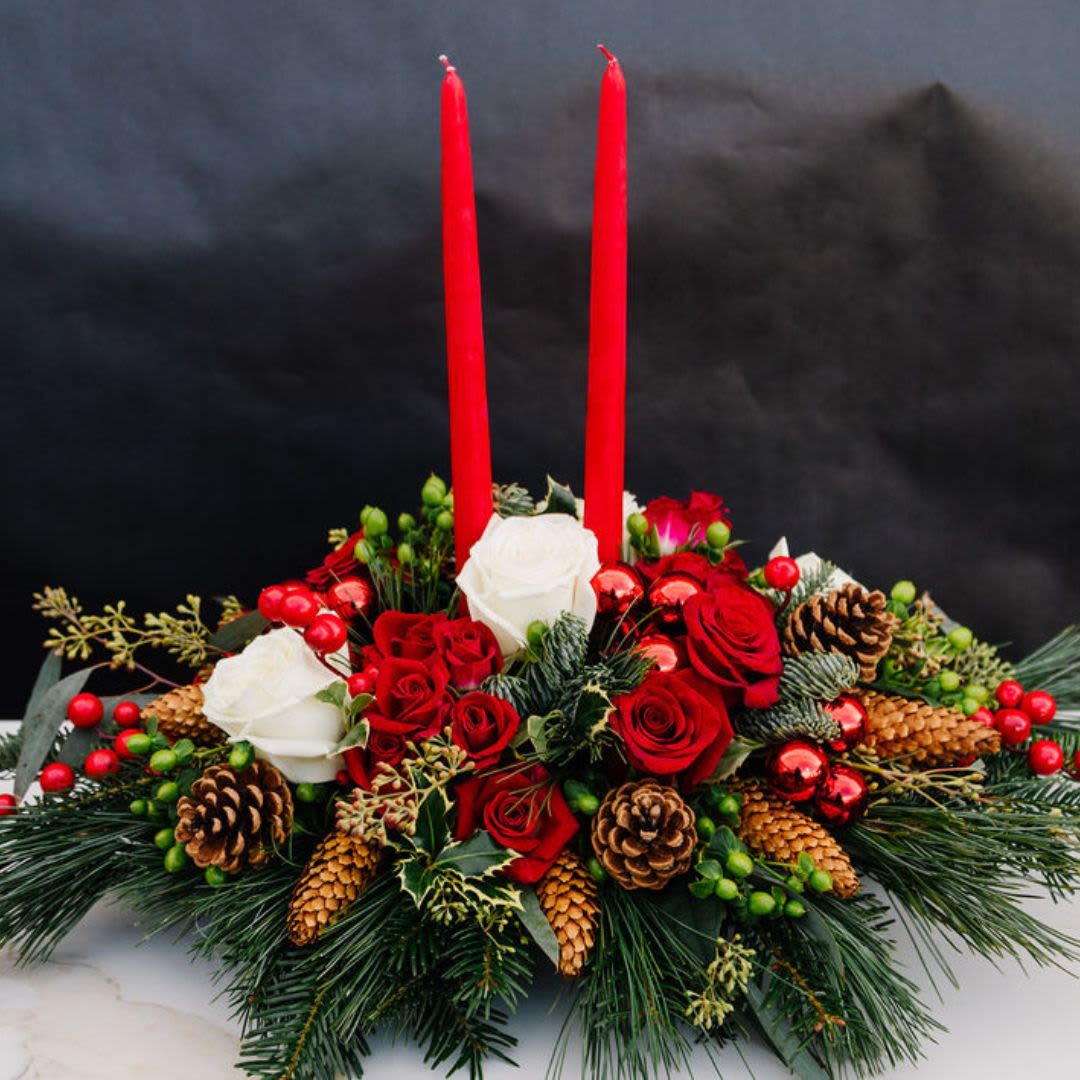 Candlelit Christmas Splendor - A timeless Christmas centerpiece featuring vibrant red roses, classic white roses, and festive red berries, accented by fresh greenery and holly. Pine cones and bright green accents add a rustic charm, while two tall red taper candles create an elegant and warm ambiance. This lush arrangement is perfect for gracing a holiday dinner table or adding a cozy, festive touch to any celebration.