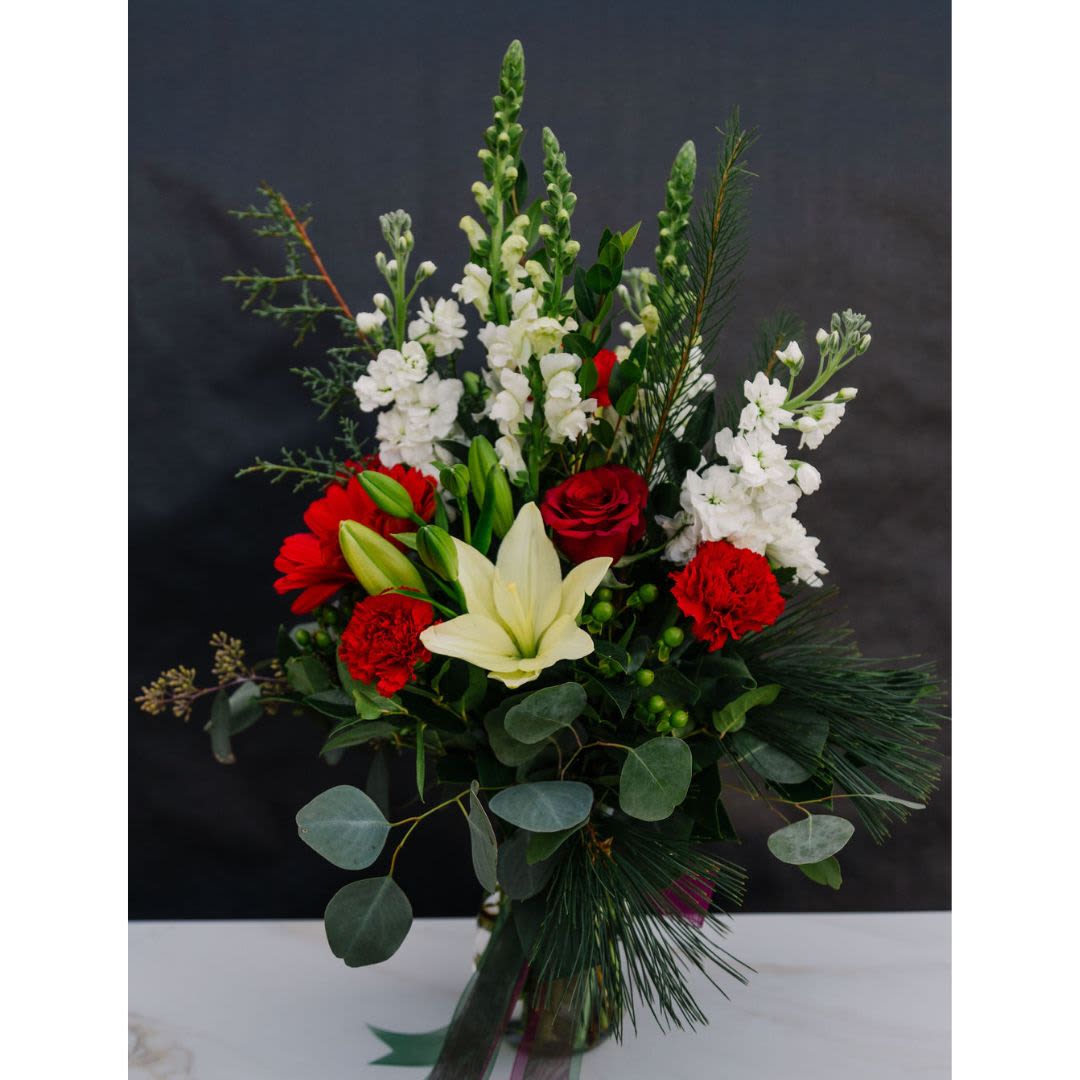 Christmas Classic - A striking arrangement blending the bold hues of red roses, carnations, and gerbera daisies with the pristine white of snapdragons and stock. A creamy lily adds sophistication, while lush greenery, including pine sprigs and eucalyptus, brings a fresh, natural touch. Perfect for holiday celebrations or a seasonal gift that radiates warmth and cheer.
