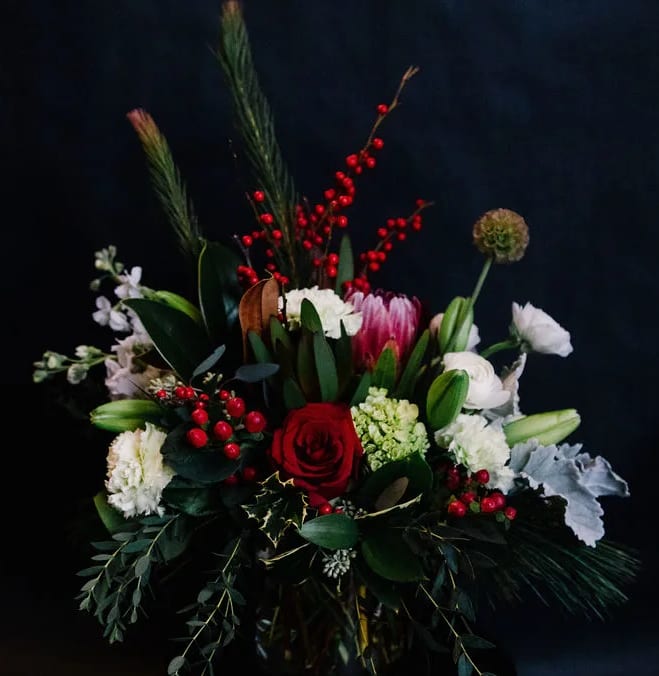 Festive Dreams - A festive mix of lux holiday blooms designed in  a low cylinder.  Roses, ranunculus, hydrangea and stock are accented with pine, eucalyptus and holly.