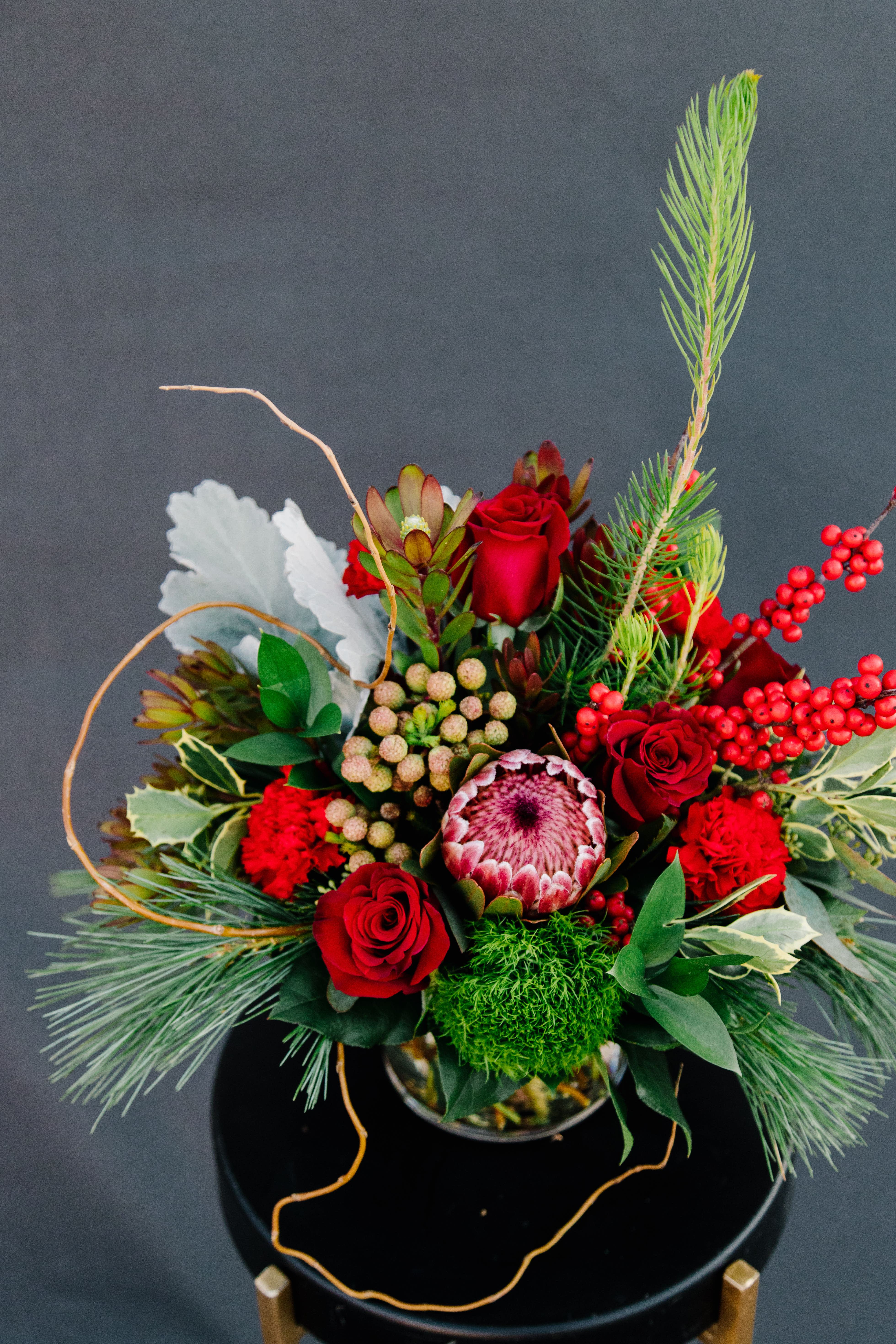 Holiday Shine - Designed in a low modern cylinder, this holiday arrangement includes a unique assortment of blooms, winter foliage, berries and pine.