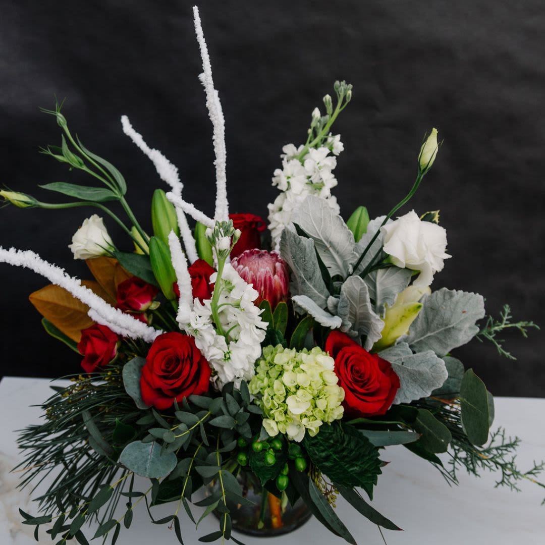 Winter's Embrace - This stunning holiday arrangement features a rich blend of seasonal blooms and greenery, designed to evoke a sense of warmth and festive cheer. The combination of rich hues, soft textures, and festive elements makes &quot;Winter's Embrace&quot; a perfect holiday piece, radiating both elegance and warmth.