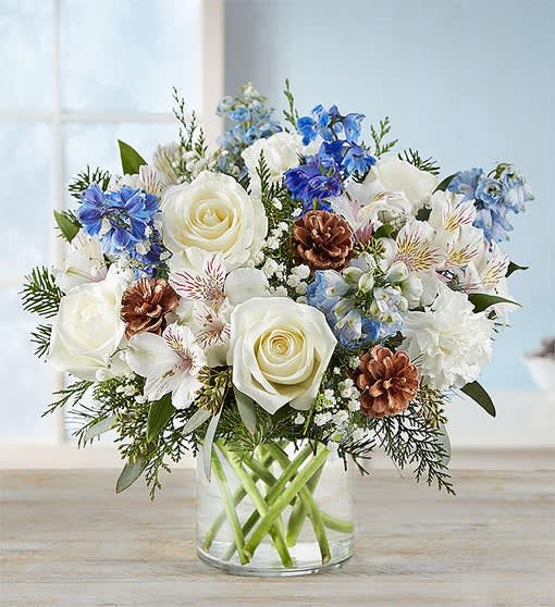 Winter Wishes Bouquet - This  arrangement with white roses, carnations, and Peruvian lilies (alstroemeria); blue delphinium; accented with baby’s breath, seeded eucalyptus, assorted Christmas greenery, and pinecones
