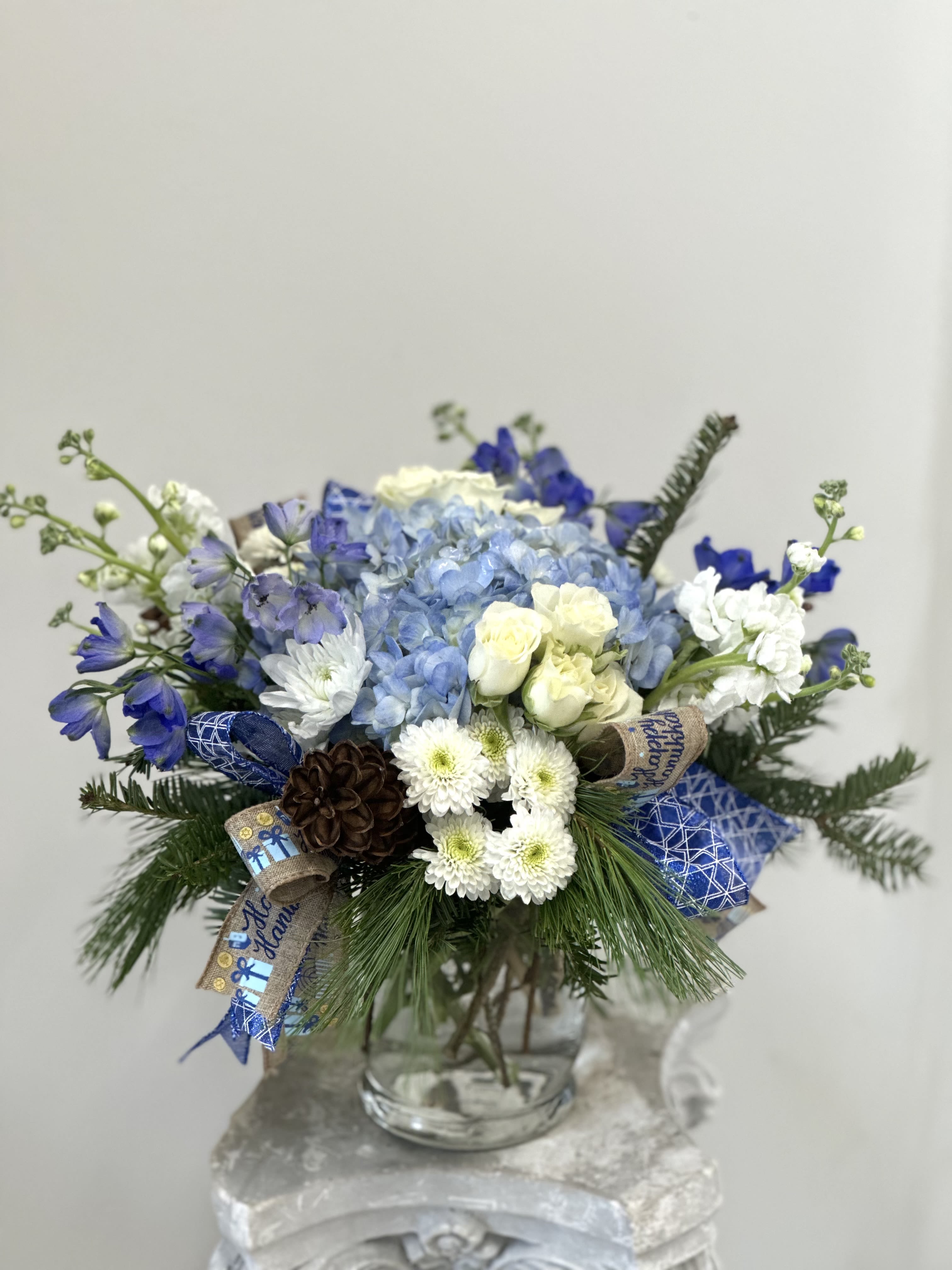 Hanukkah Harmony by Westford Florist - Celebrate the magic of Hanukkah with our enchanting arrangement, Hanukkah Harmony by Westford Florist! This stunning bouquet features delicate blue hydrangeas dancing alongside elegant spray roses and vibrant delphinium. Each bloom is artfully nestled among fluffy white stock, charming white poms, and whimsical dot poms—perfectly complemented by natural pine cones for a touch of festive warmth. Topped off with a beautiful Hanukkah ribbon, this floral masterpiece transforms any space into a winter wonderland. Bring joy to your celebrations or share the love with family and friends—Hanukkah Harmony is sure to light up every heart!
